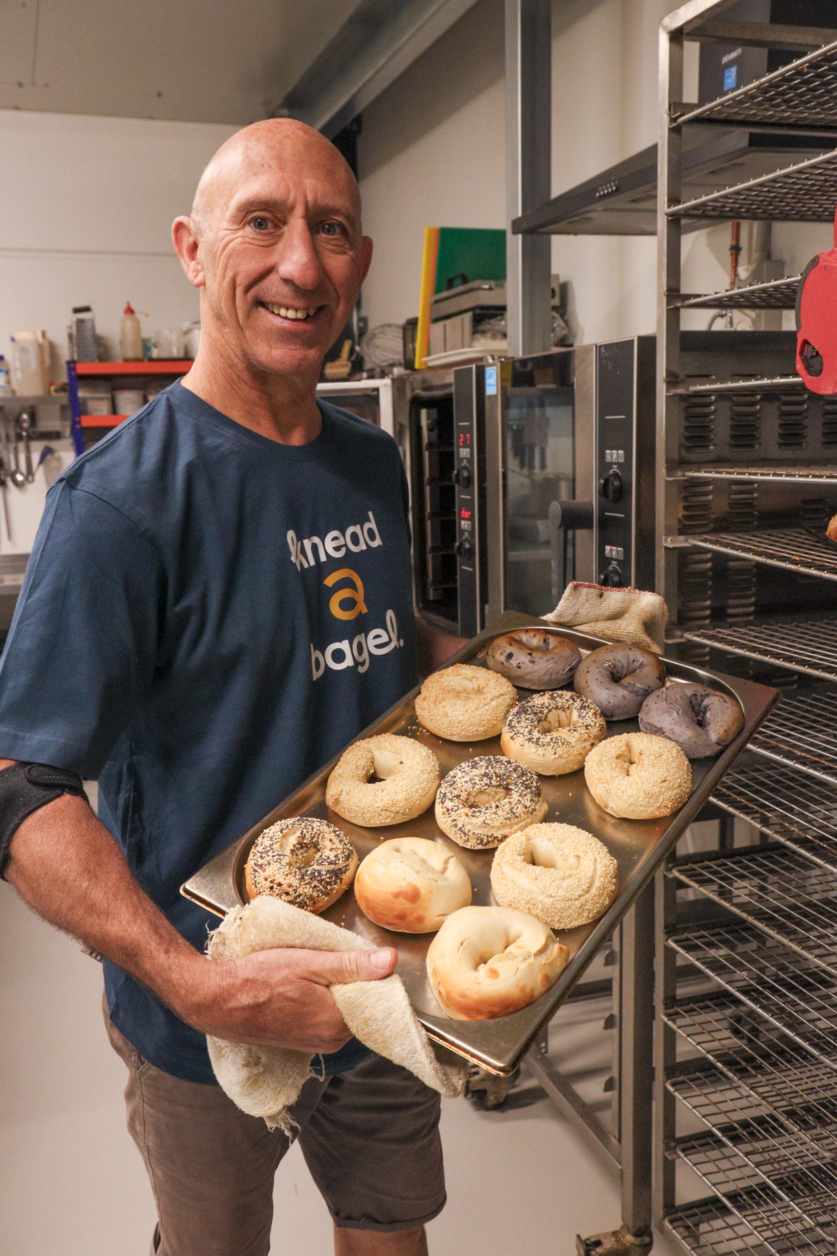 A smiling man with a shaved head holding a tray of assorted donuts in a commercial kitchen or bakery setting.
