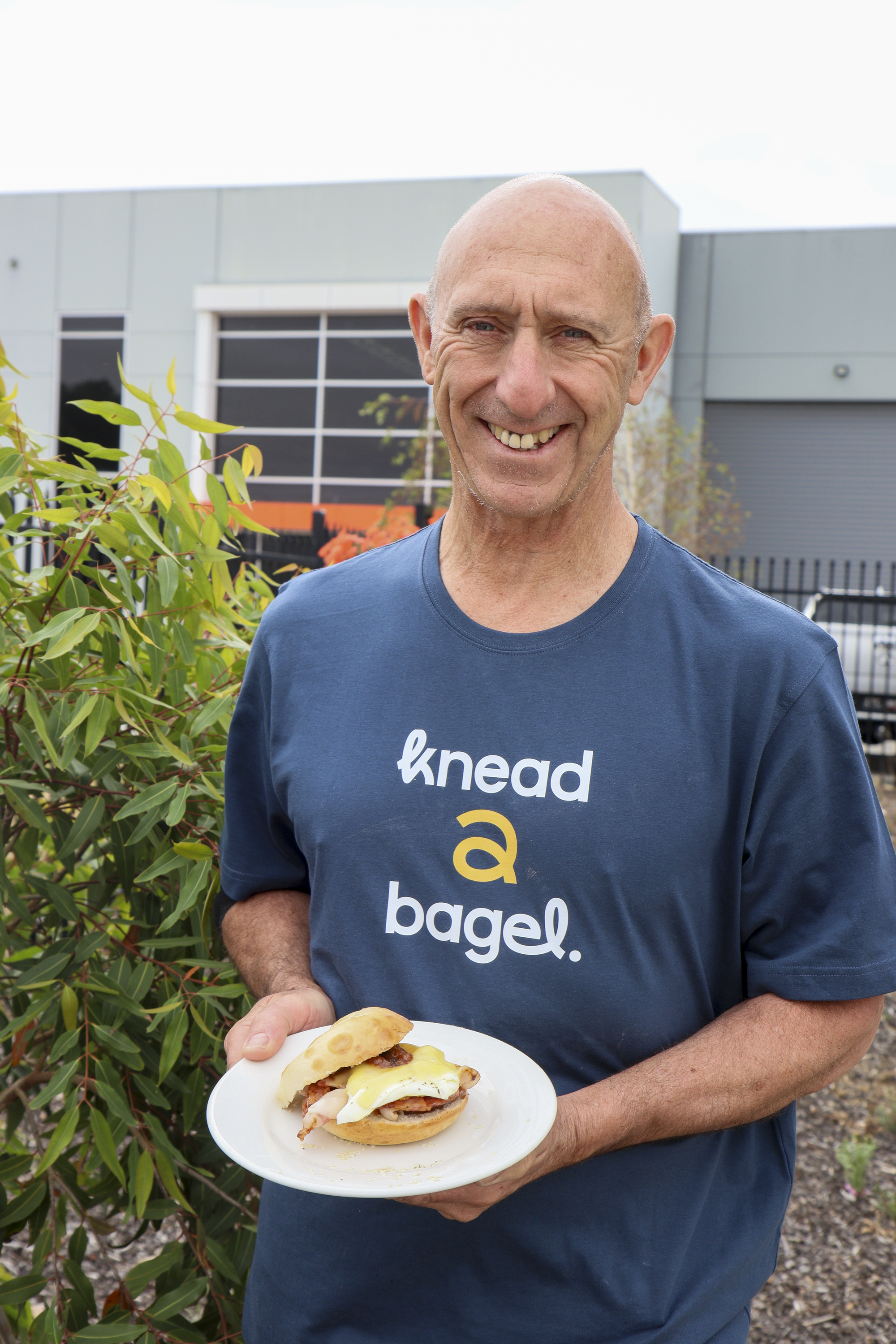 A smiling man wearing a blue t-shirt holding a plate with a breakfast sandwich, standing outdoors near green bushes and an industrial building in the background.