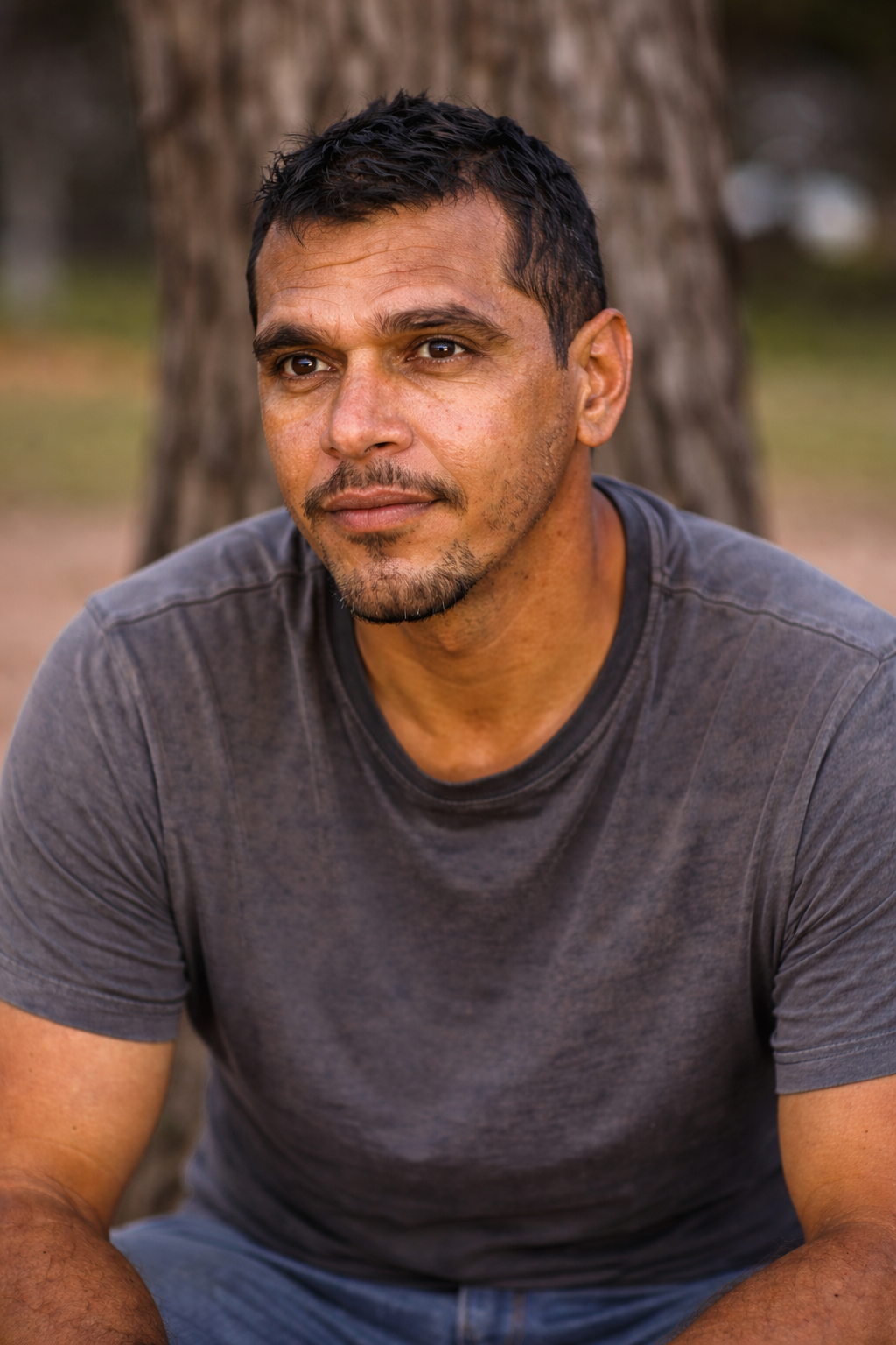 A man with short dark hair and a beard, wearing a gray t-shirt, sits outdoors near a tree trunk, looking contemplative.