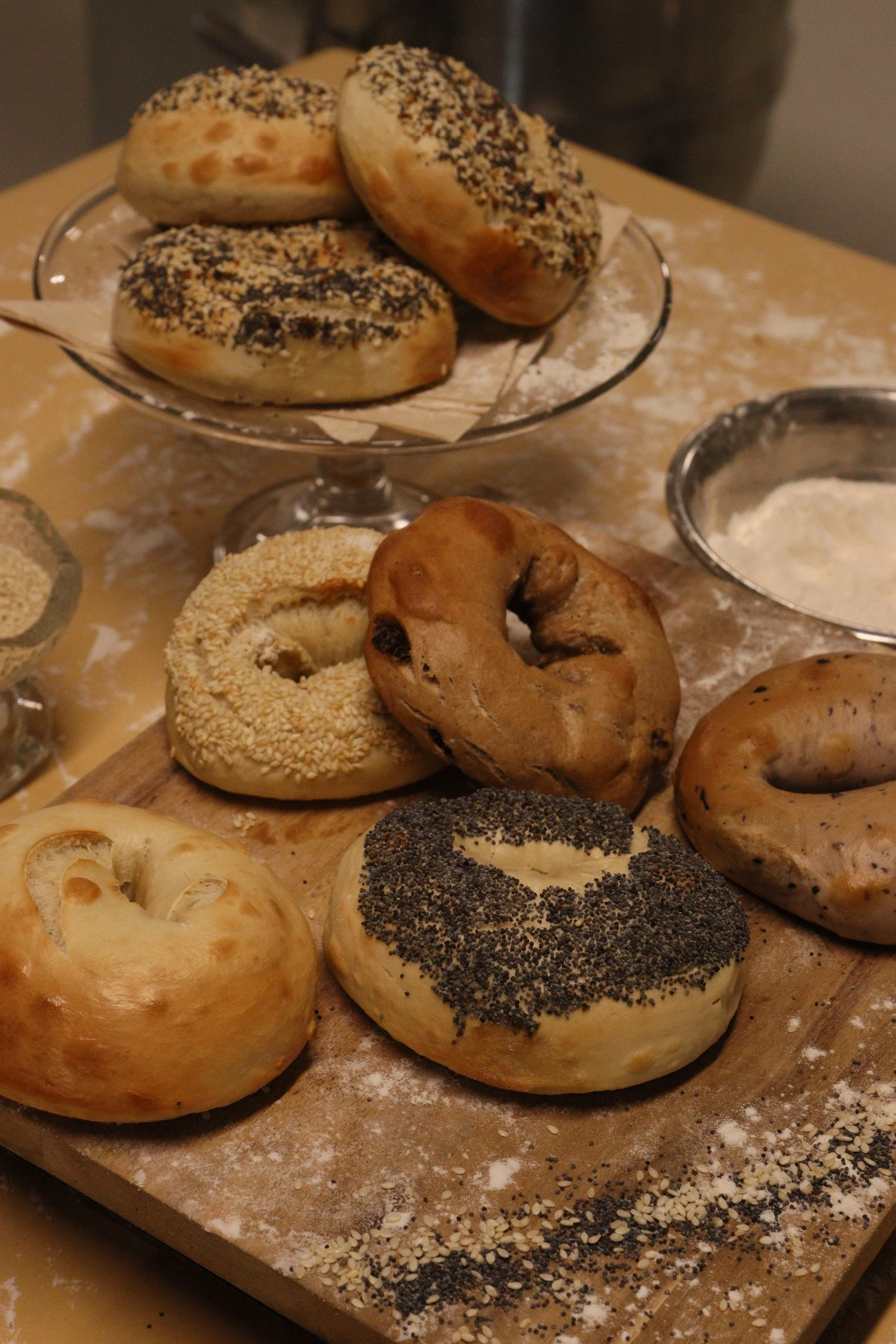 Assorted bagels with toppings such as sesame seeds, poppy seeds, and chopped nuts on a wooden cutting board, with flour and seeds scattered around.