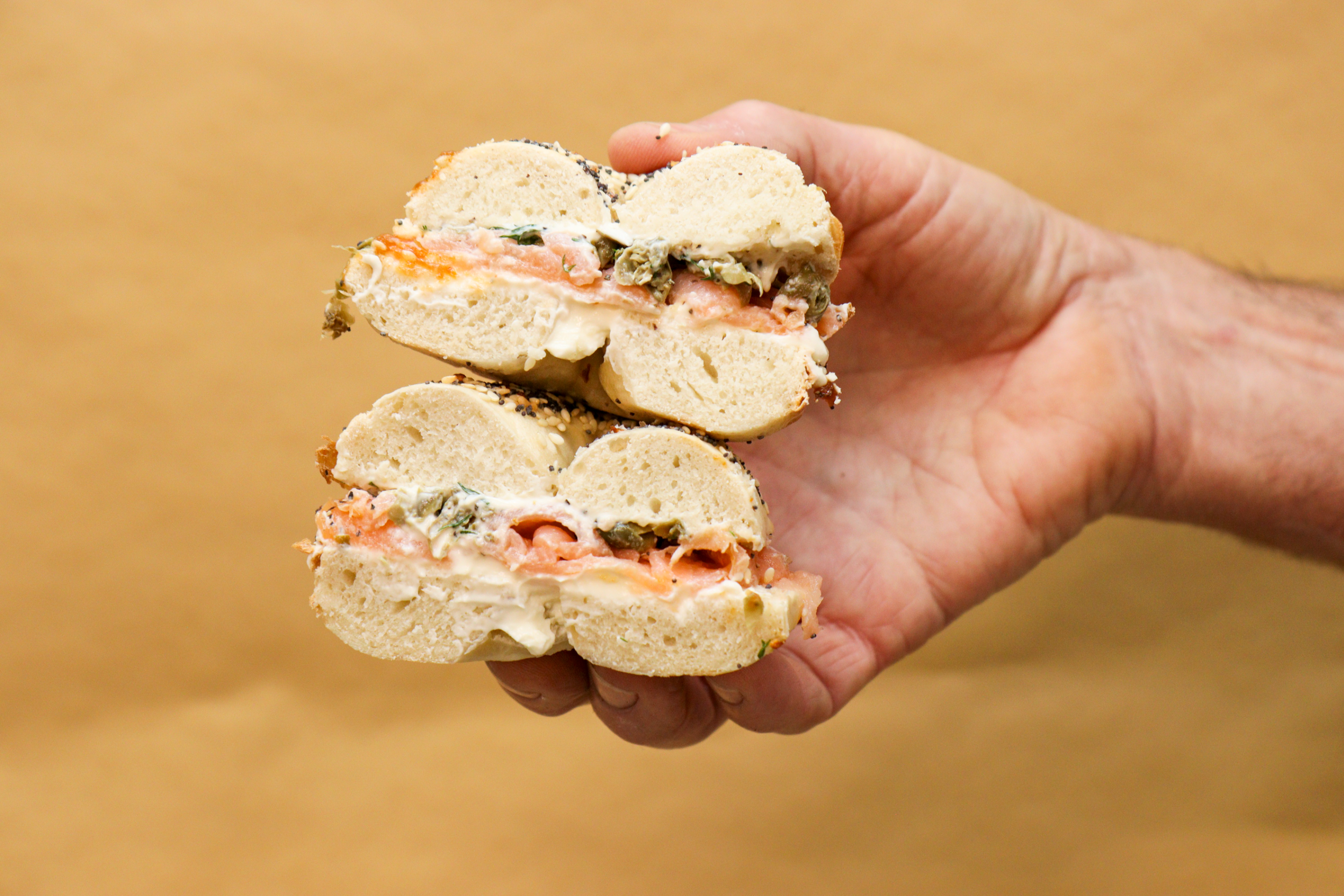 Close-up of a hand holding a broken bagel sandwich with cream cheese, salmon, and herbs.