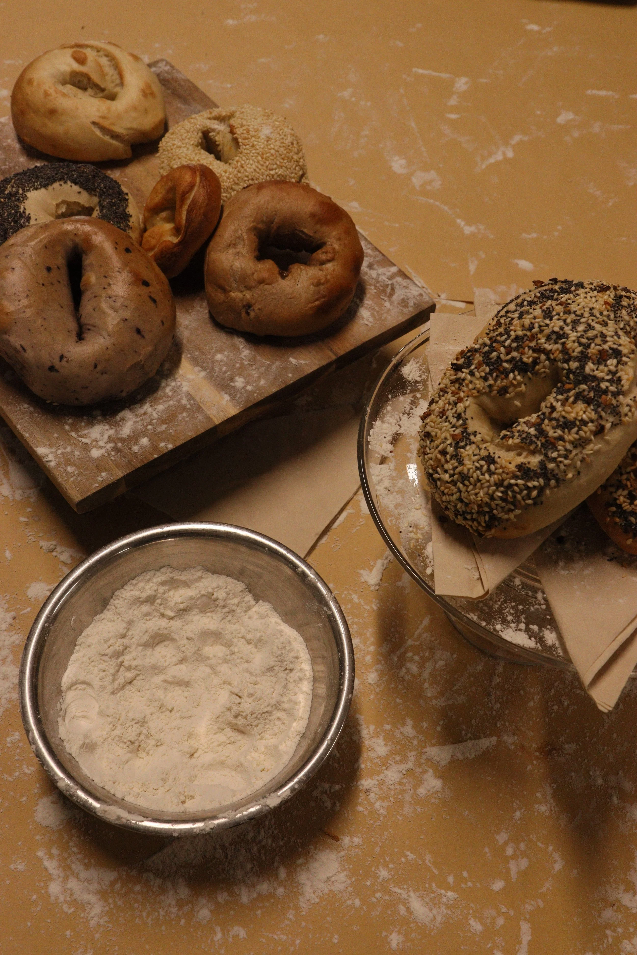 Assorted bagels on a wooden cutting board, a bagel with sesame seeds, a bagel with poppy seeds, and others, with a bowl of flour and a rolled-out dough sheet on a floured surface.