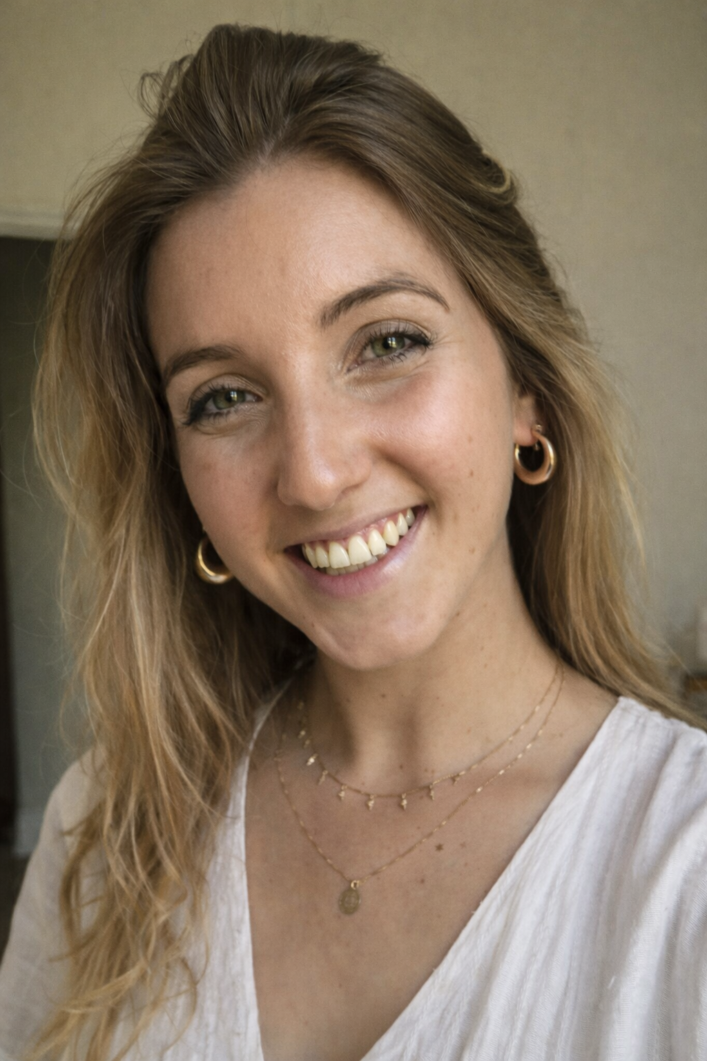 A young woman with wavy blonde hair smiling, wearing gold hoop earrings, layered gold necklaces, and a white top.