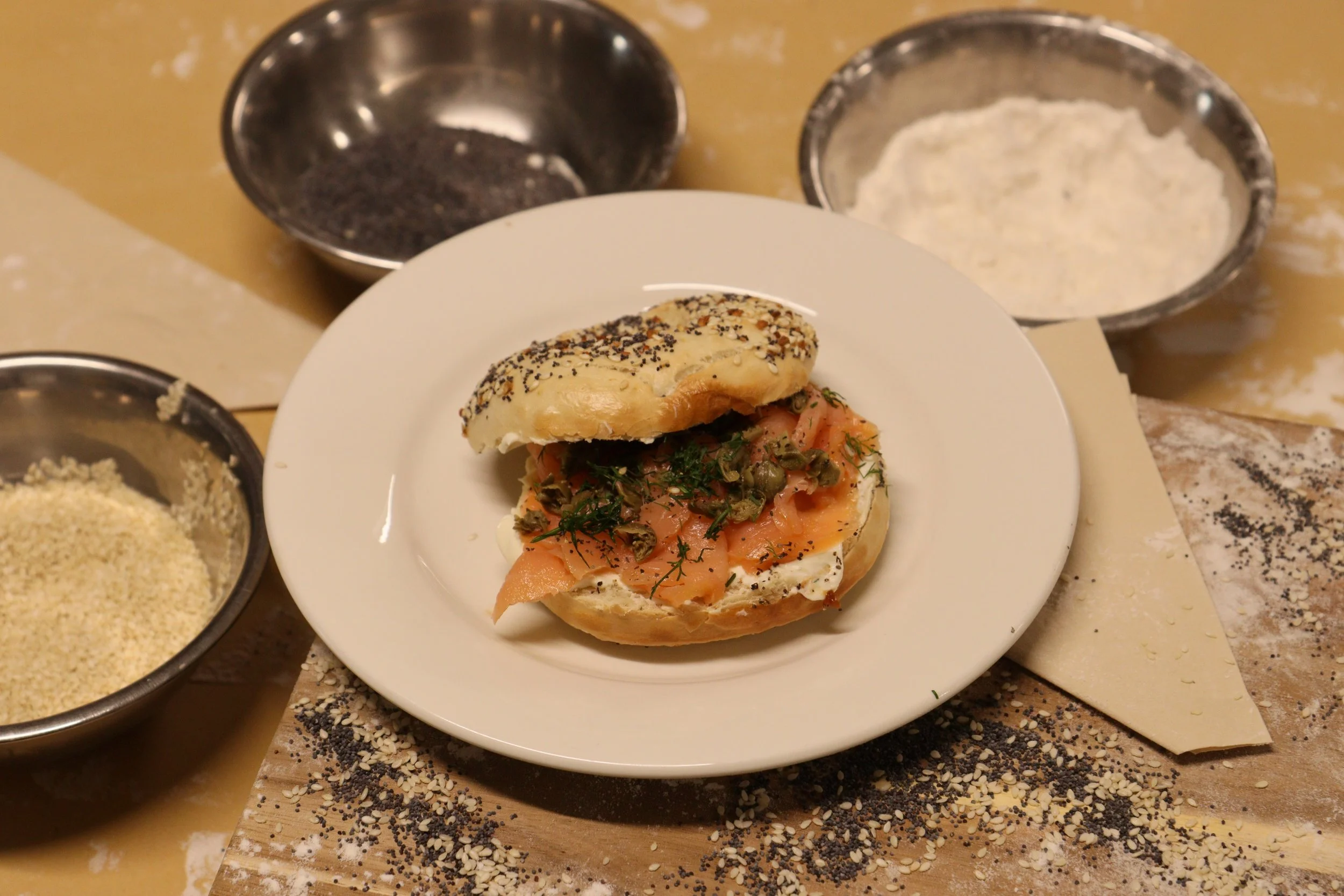 A sandwich with smoked salmon, cream cheese, and herbs on a sesame seed bun, placed on a white plate. Surrounding it are bowls of poppy seeds, sesame seeds, and flour, with a wooden cutting board covered in sesame seeds and poppy seeds.