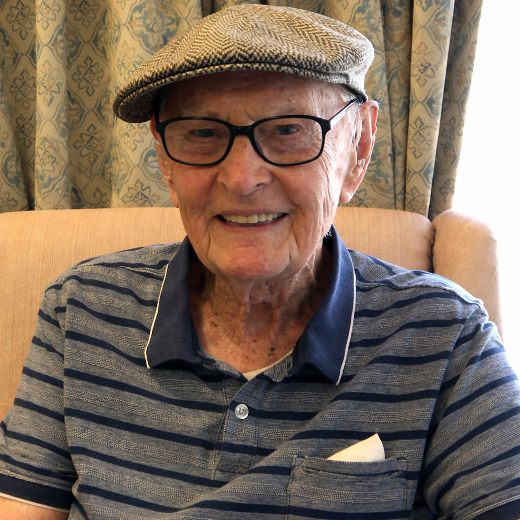 An elderly man with glasses, a patterned cap, and a striped polo shirt smiling while sitting on a beige chair in front of a patterned curtain.