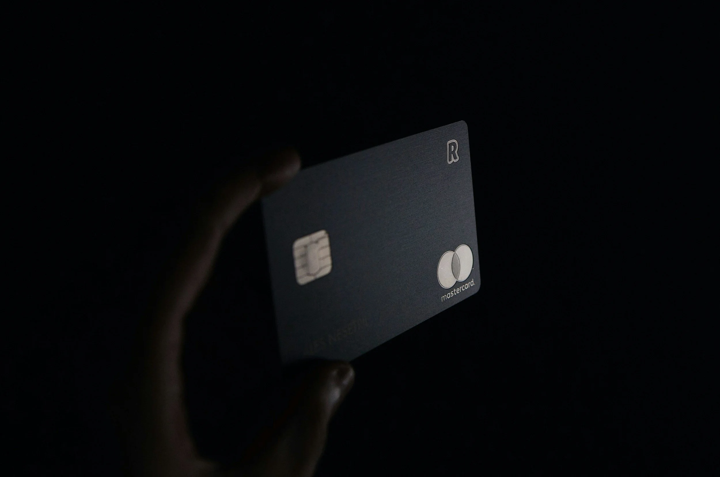 A hand holding a dark gray Mastercard credit card in a dark environment.