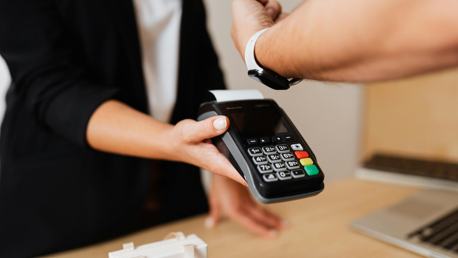 A person making a contactless payment with a smart watch using a Point of Sale terminal, while holding the terminal with their other hand.