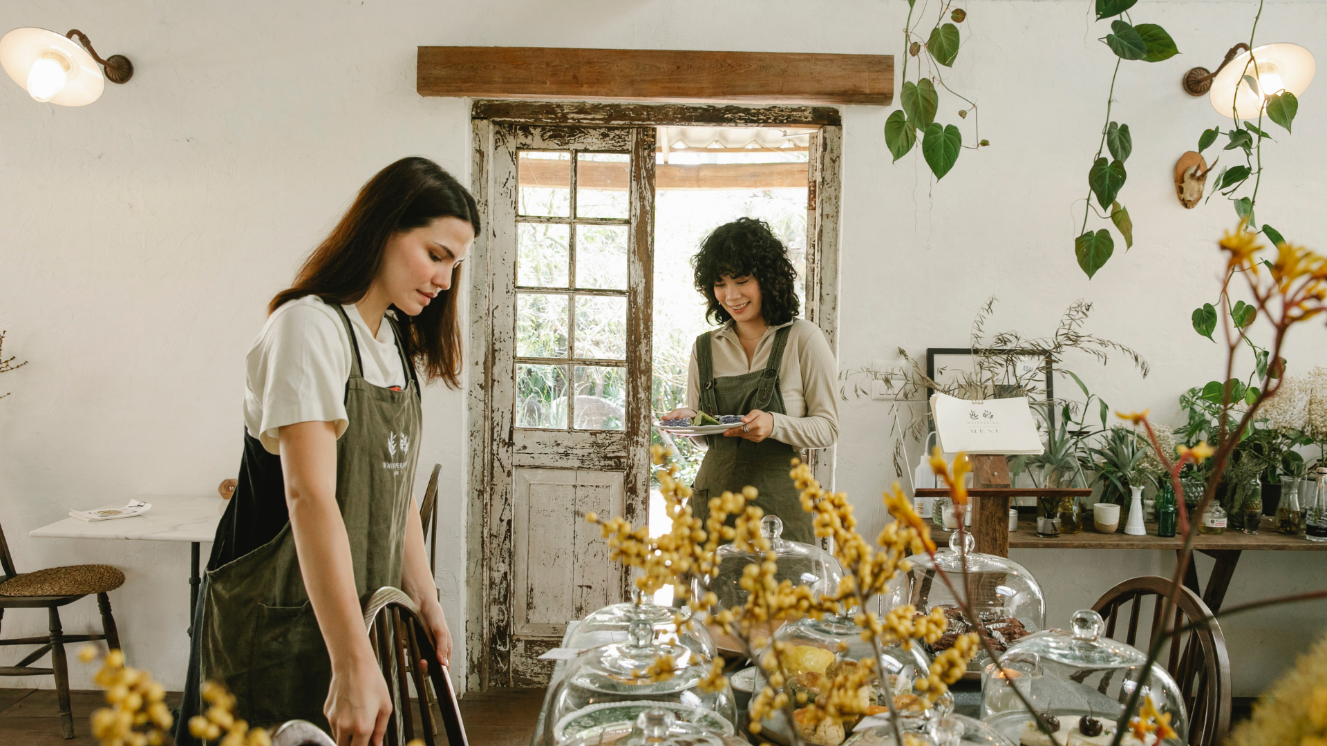 Two women in aprons setting a table with desserts in a cozy, rustic cafe with plants and natural decor.