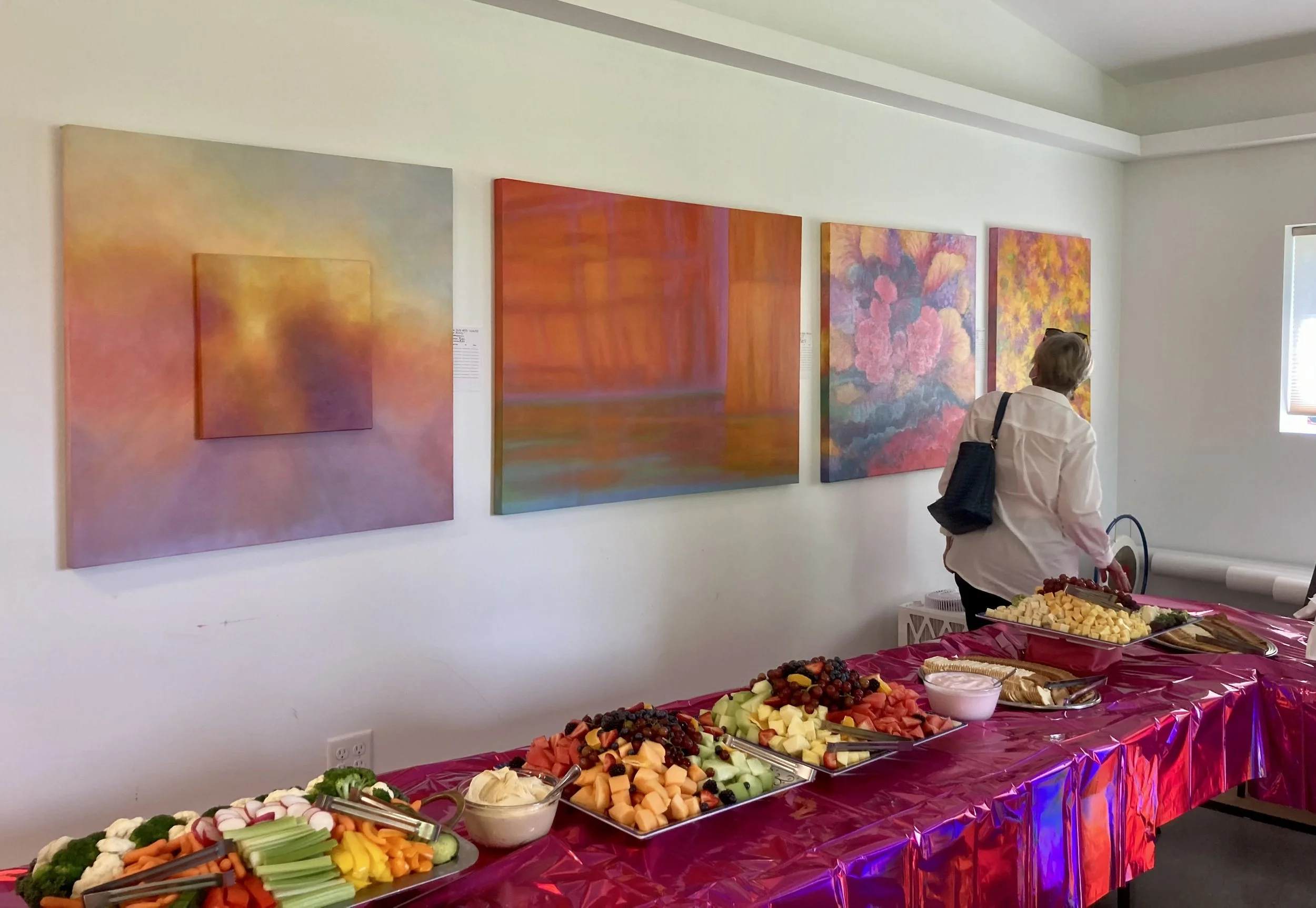 A woman is standing near a table with an assortment of fresh fruit, cheese, and snacks at an art gallery, with colorful abstract paintings hanging on the wall behind her.