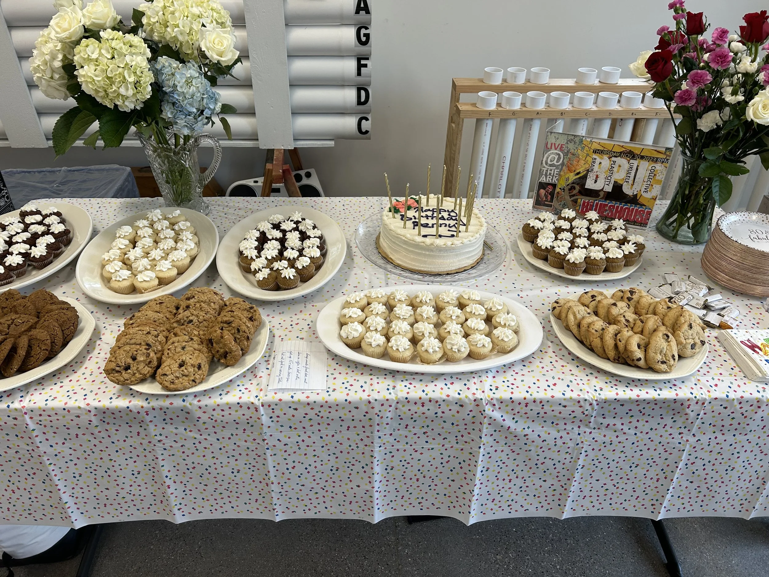 Party table with cupcakes, cookies, a birthday cake with candles, and floral arrangements.