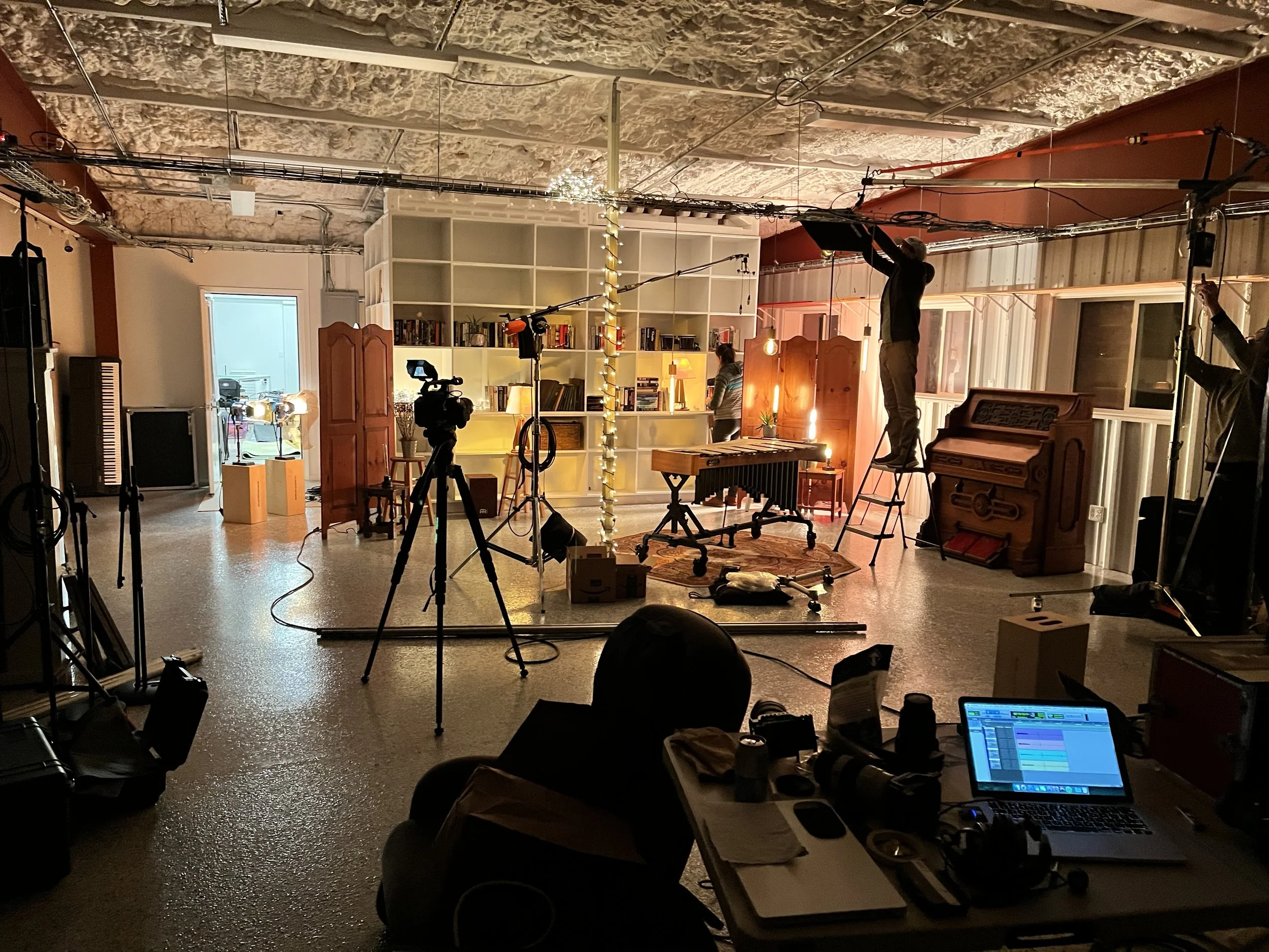 Inside a film or photo set with crew members setting up equipment and lighting, a camera on a tripod, and a person standing on a ladder adjusting lights in a cozy room with a bookshelf, piano, and decorative items.