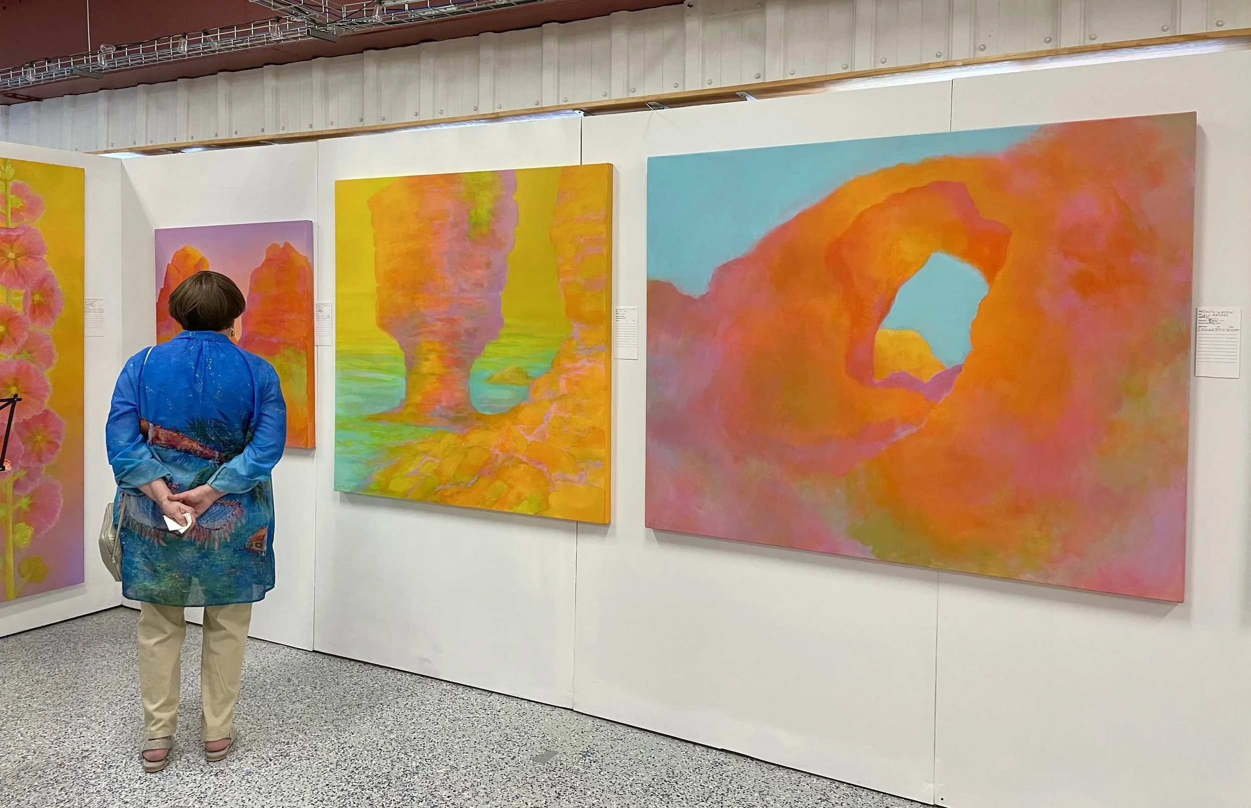 A woman with short brown hair, wearing a blue jacket and khaki pants, stands with her hands behind her back, observing vibrant abstract paintings on a white gallery wall. The paintings feature bright colors and resemble landscapes with rock formations and sky elements.