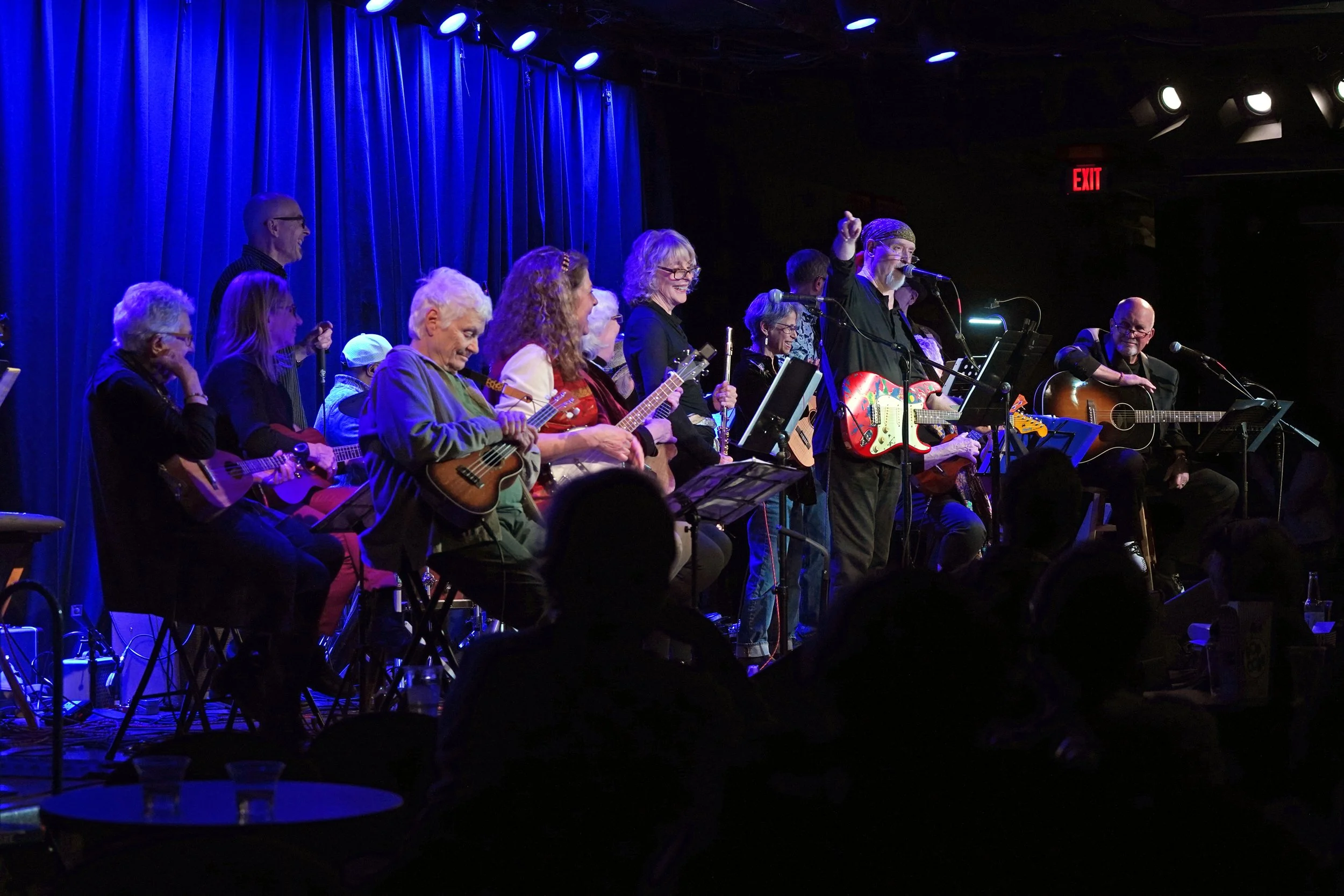 A group of musicians playing guitars and ukuleles on a stage with blue curtains and stage lighting, performing for an audience.