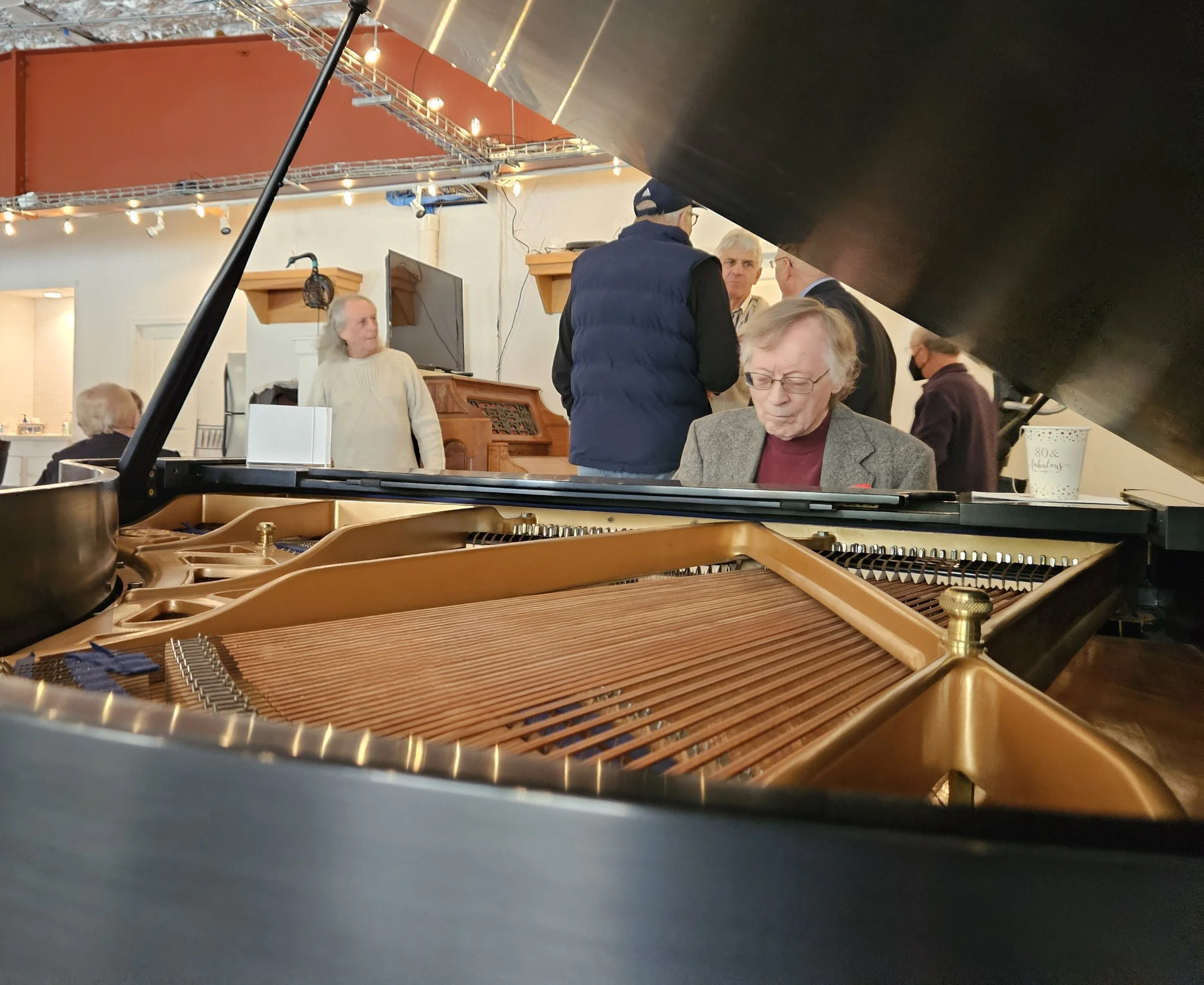 View of the inside of a grand piano with people gathered in the background, including an elderly man seated, a woman wearing a cream sweater, and a group of people standing and talking.