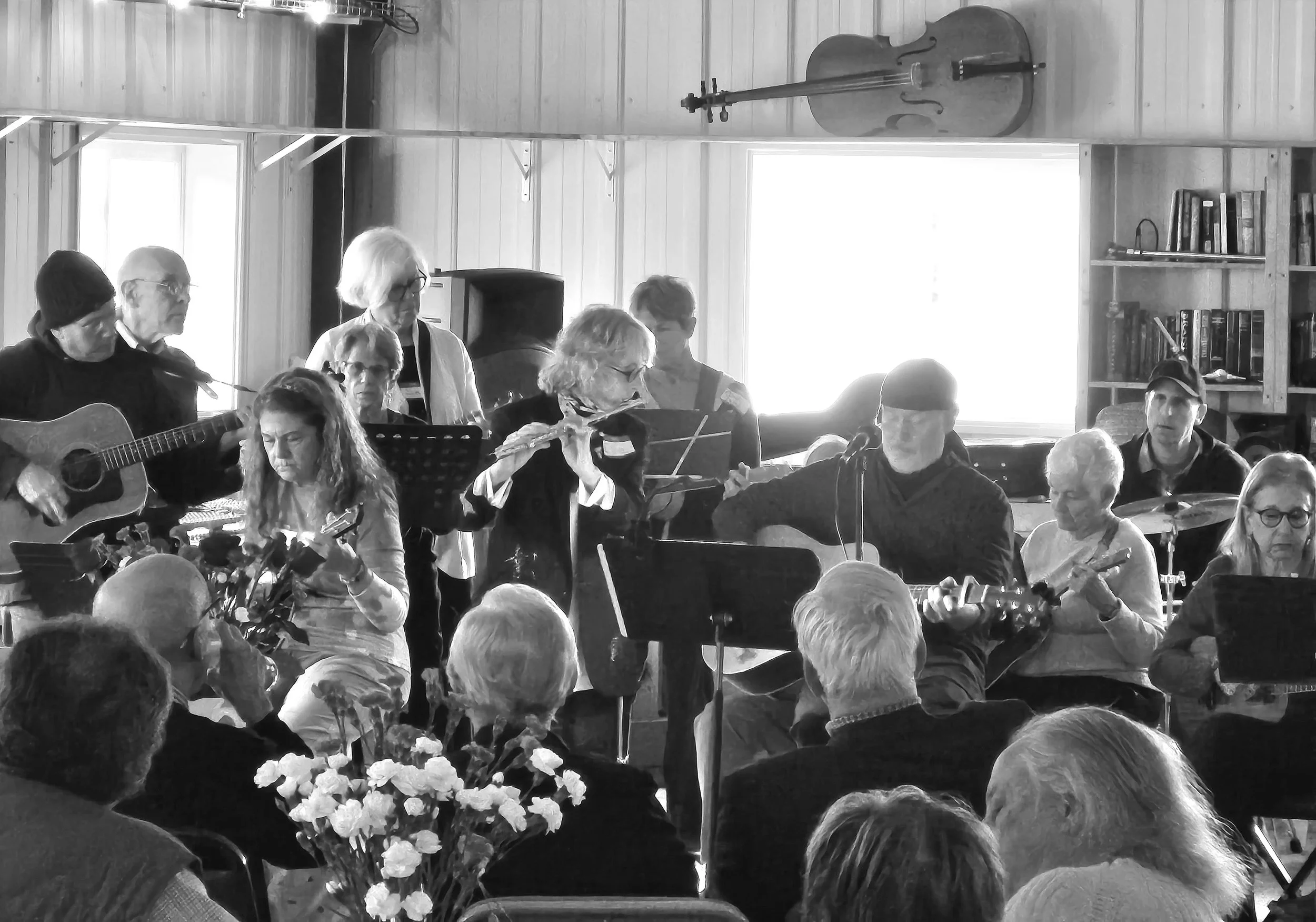 A group of elderly musicians performing with guitars, a flute, and other instruments in a cozy wooden room with an audience watching, flowers on a table in foreground, books on shelves, and a double bass hanging on the wall.