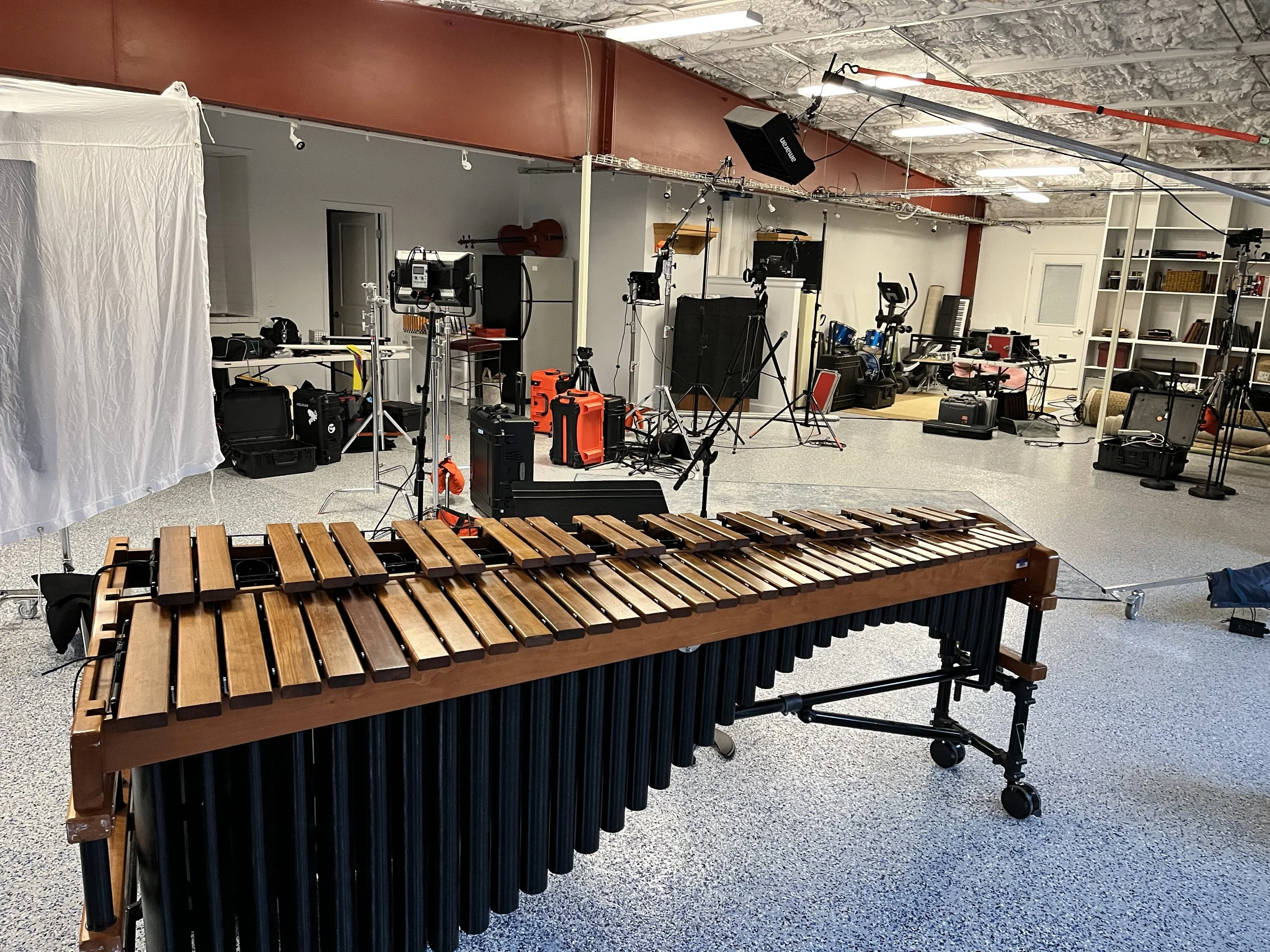 A music studio setup with a xylophone in the foreground and various musical instruments, tripods, and equipment in the background. The studio has a gray carpet, white walls, and an exposed ceiling.