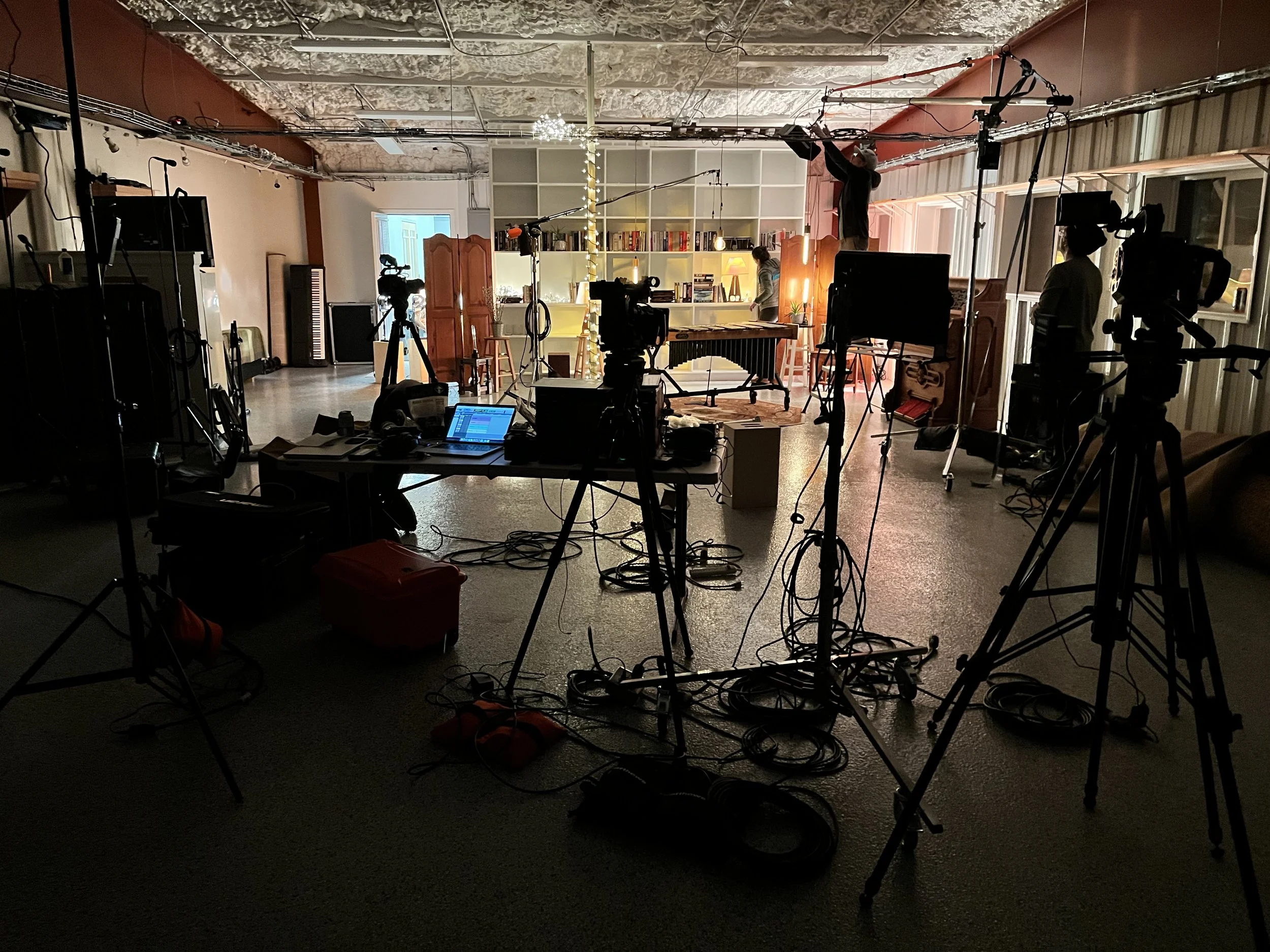 A film or photo shoot in progress with multiple cameras, tripods, lighting equipment, and crew members in a spacious room with furniture and shelves on the walls.