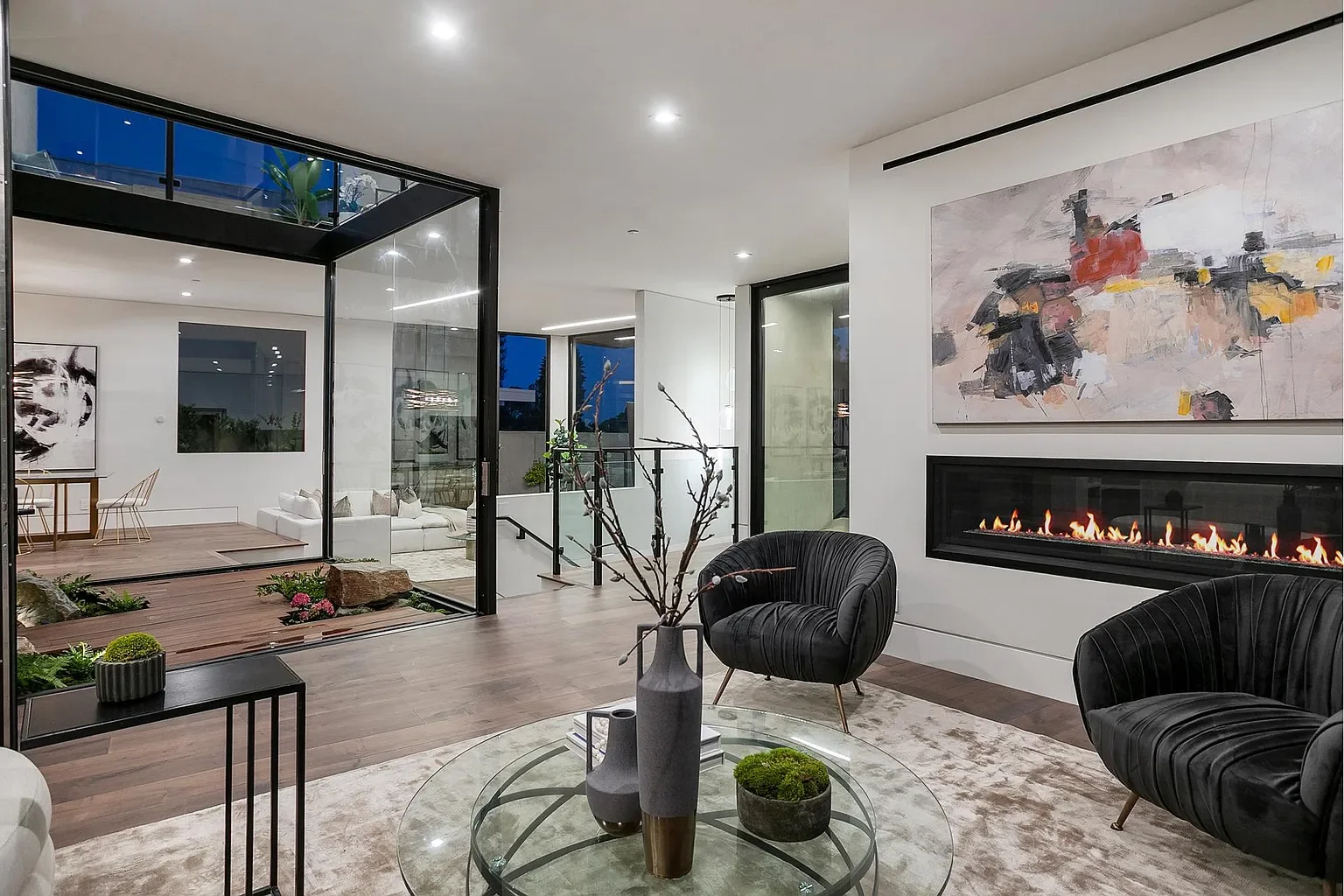 Modern living room with black leather chairs, a fireplace, abstract painting, glass coffee table with vases, and an indoor garden visible through a glass sliding door.