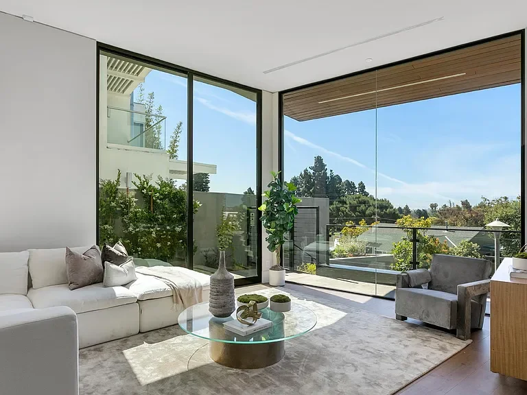 Modern living room with large floor-to-ceiling windows, a white sofa with pillows, a glass coffee table with decorative items, a gray armchair, and a view of trees and sky outside.