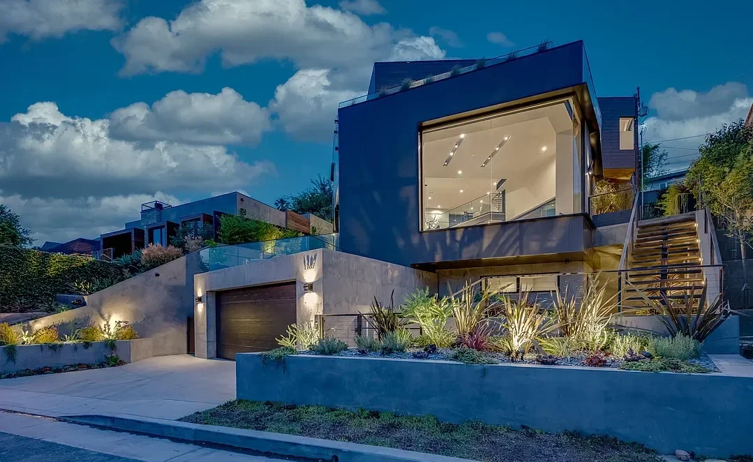 Modern multi-story house with large glass windows, concrete walls, and landscaped front yard, illuminated at dusk.