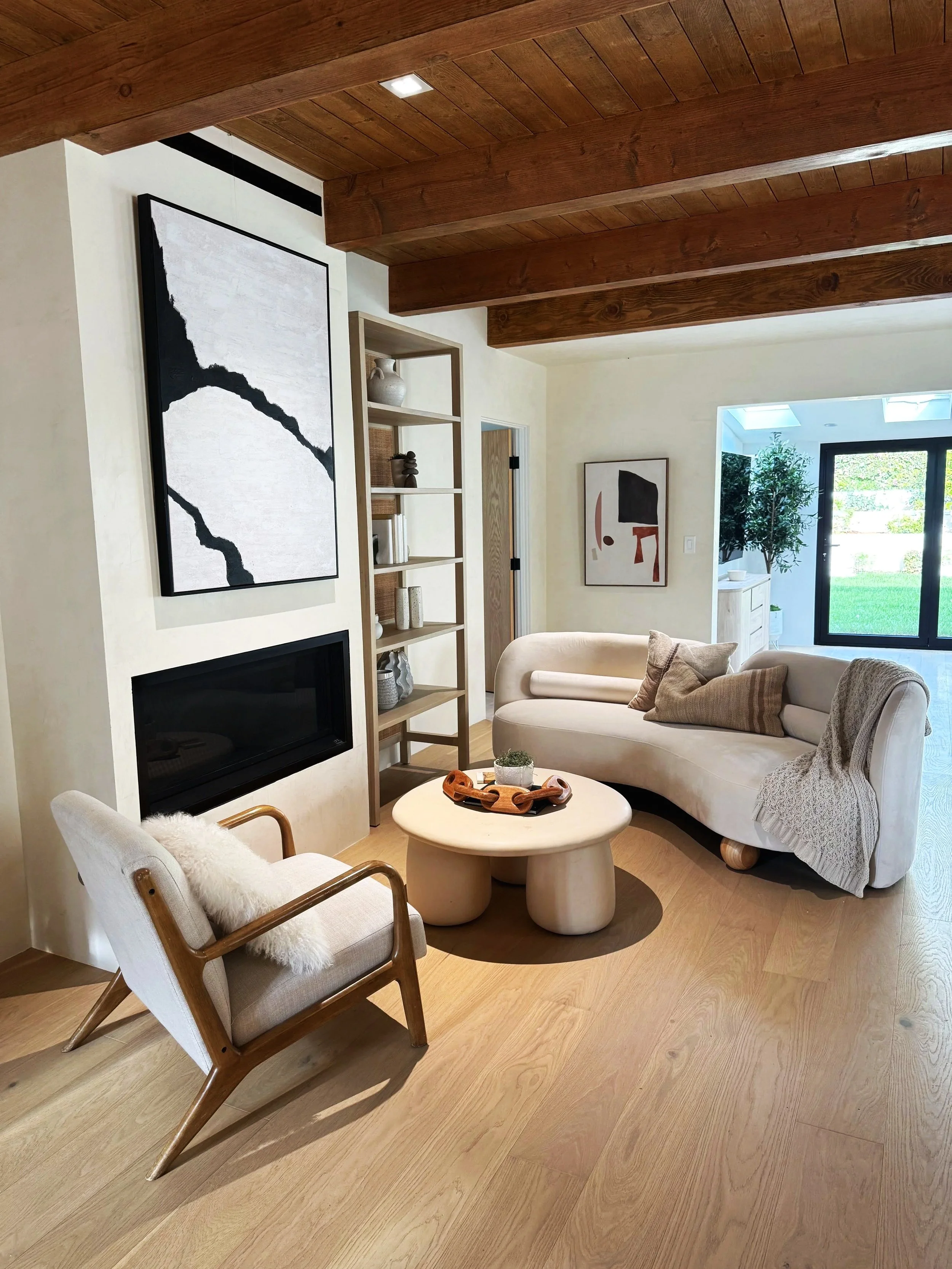 A cozy living room with light-colored furniture, a wooden ceiling with exposed beams, and a large window revealing a green outdoor space. Features include a cream curved sofa with pillows, a small round coffee table with a plant, a wooden chair with a fur throw, a modern painting on the wall, and a built-in fireplace.
