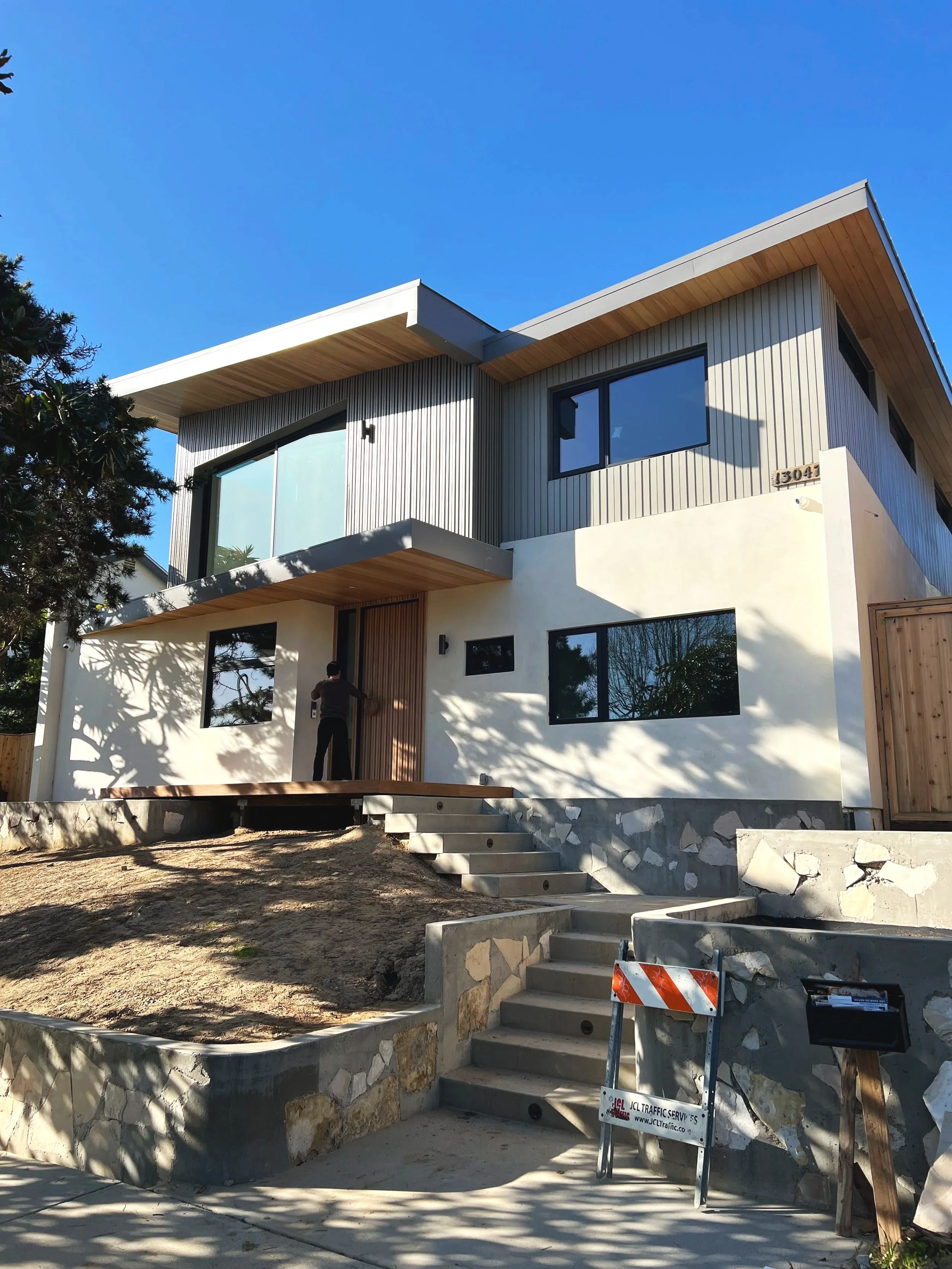 Modern two-story house under construction with a mix of white and gray exterior walls, large windows, wooden accents, and concrete stairs leading to the front door. A person is standing near the entrance, and construction equipment is visible.
