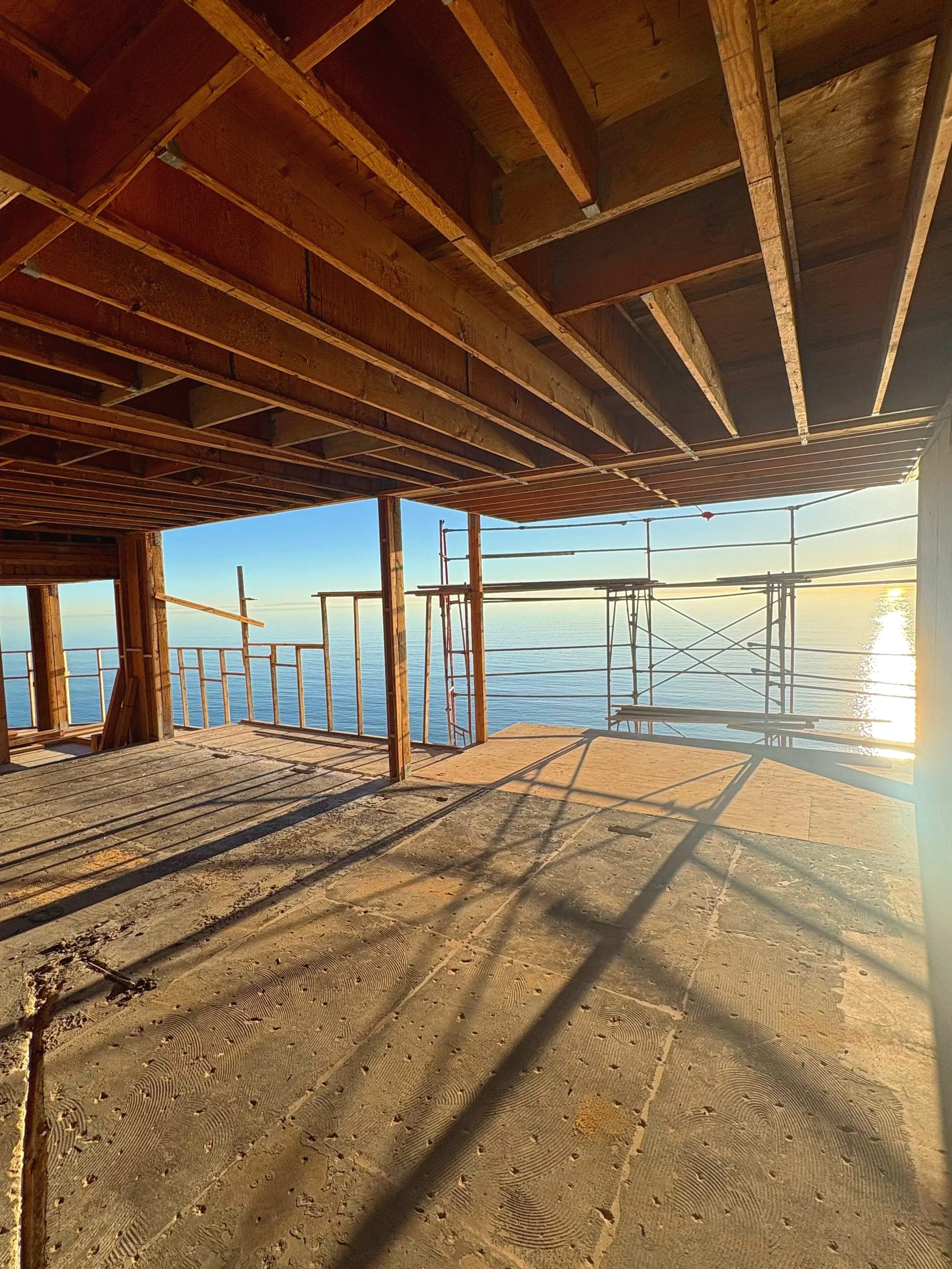 Construction site of a building with an open balcony overlooking the ocean during sunset, with scaffolding and unfinished wooden beams.