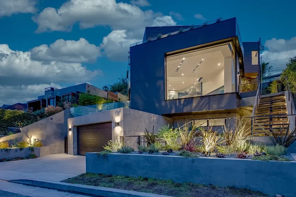 Modern house with large glass windows and exterior lighting, surrounded by landscaped plants and steps at dusk.