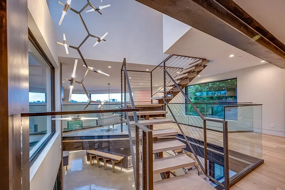 A modern interior staircase with wooden steps, metal railing, and glass panels, leading to an upper level. Large windows provide natural light and views of the outdoors. A contemporary chandelier resembling a molecular structure hangs above.