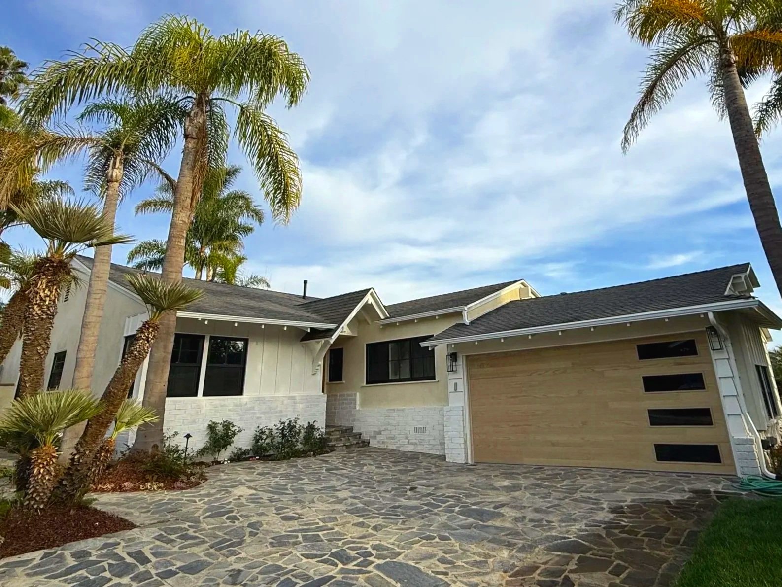 Modern house with stone pathway, palm trees, and a partly cloudy sky.