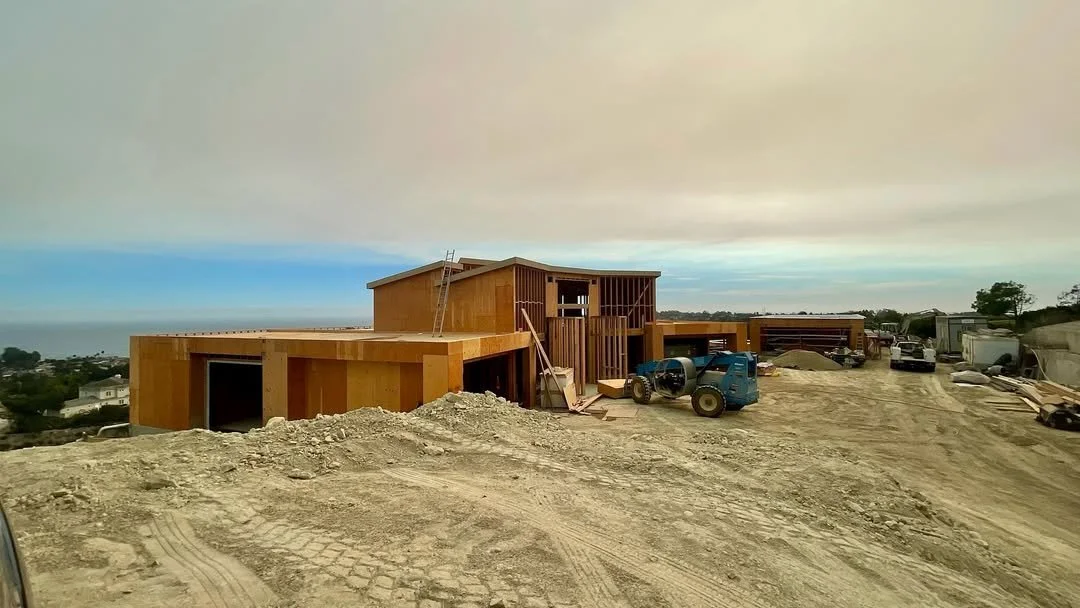 Under construction wooden house on a hill with construction equipment and vehicles nearby, overlooking a landscape with trees and houses.