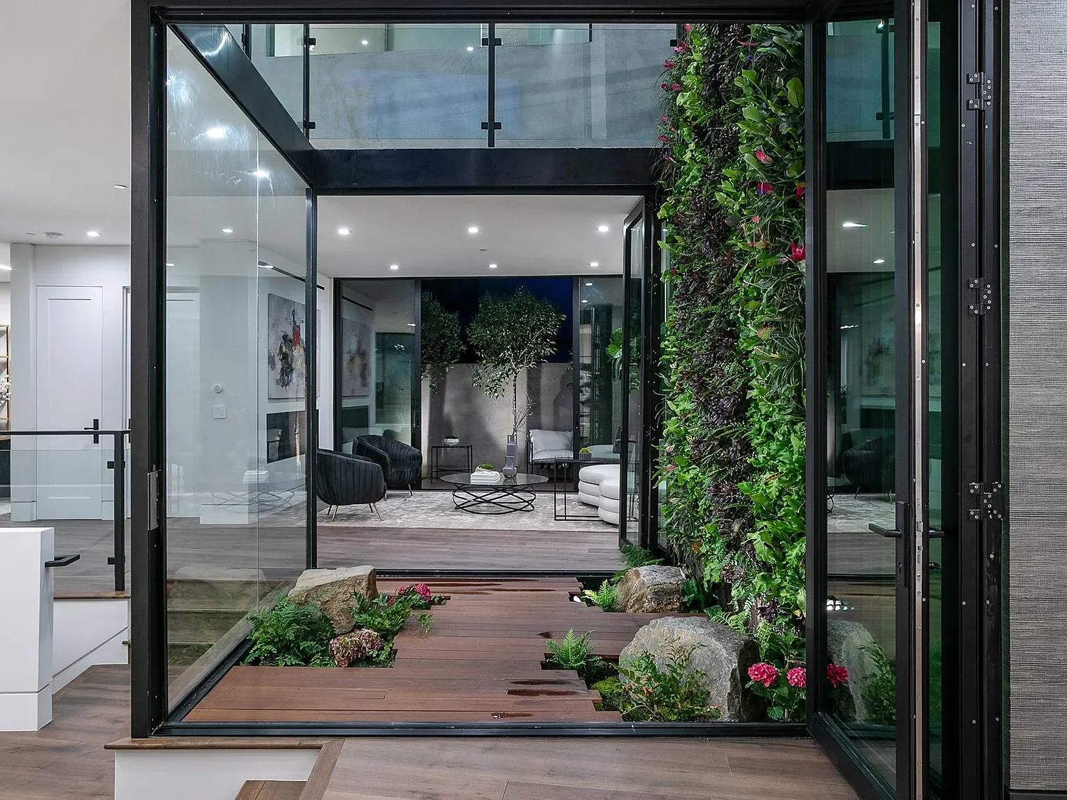 Contemporary indoor living room with large glass doors opening to an outdoor deck with a vertical garden and rocks, modern furniture, and interior artwork.