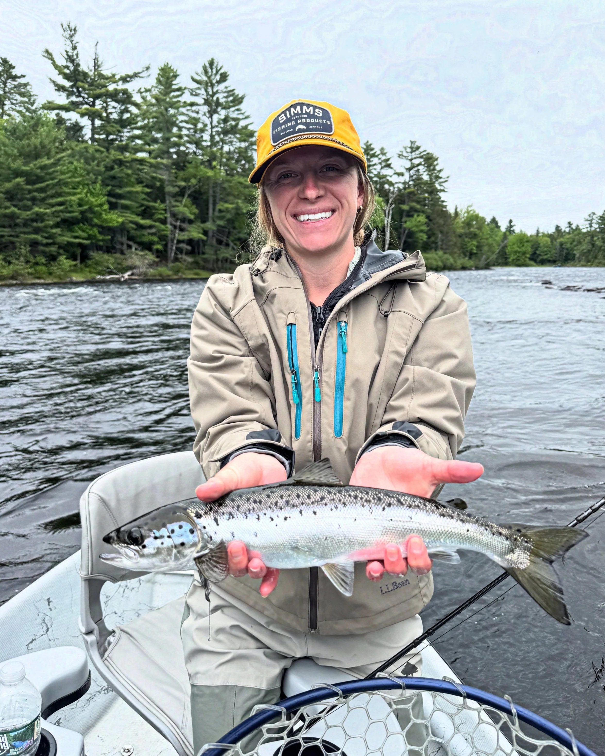 A woman on a boat holding a fish with trees and water in the background.