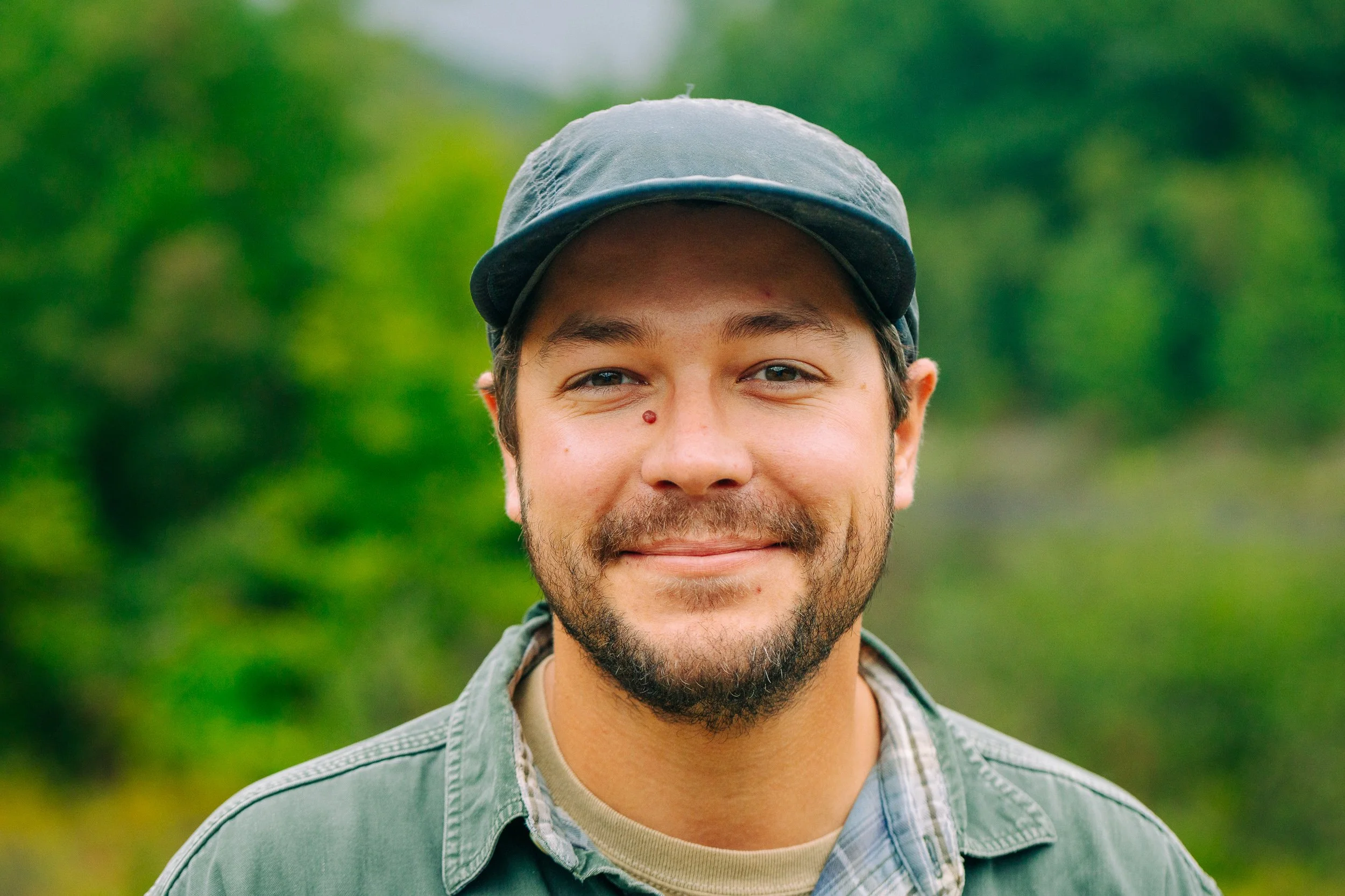 A smiling young man with a beard and mustache wearing a dark baseball cap and green jacket outdoors with blurred green foliage in the background.