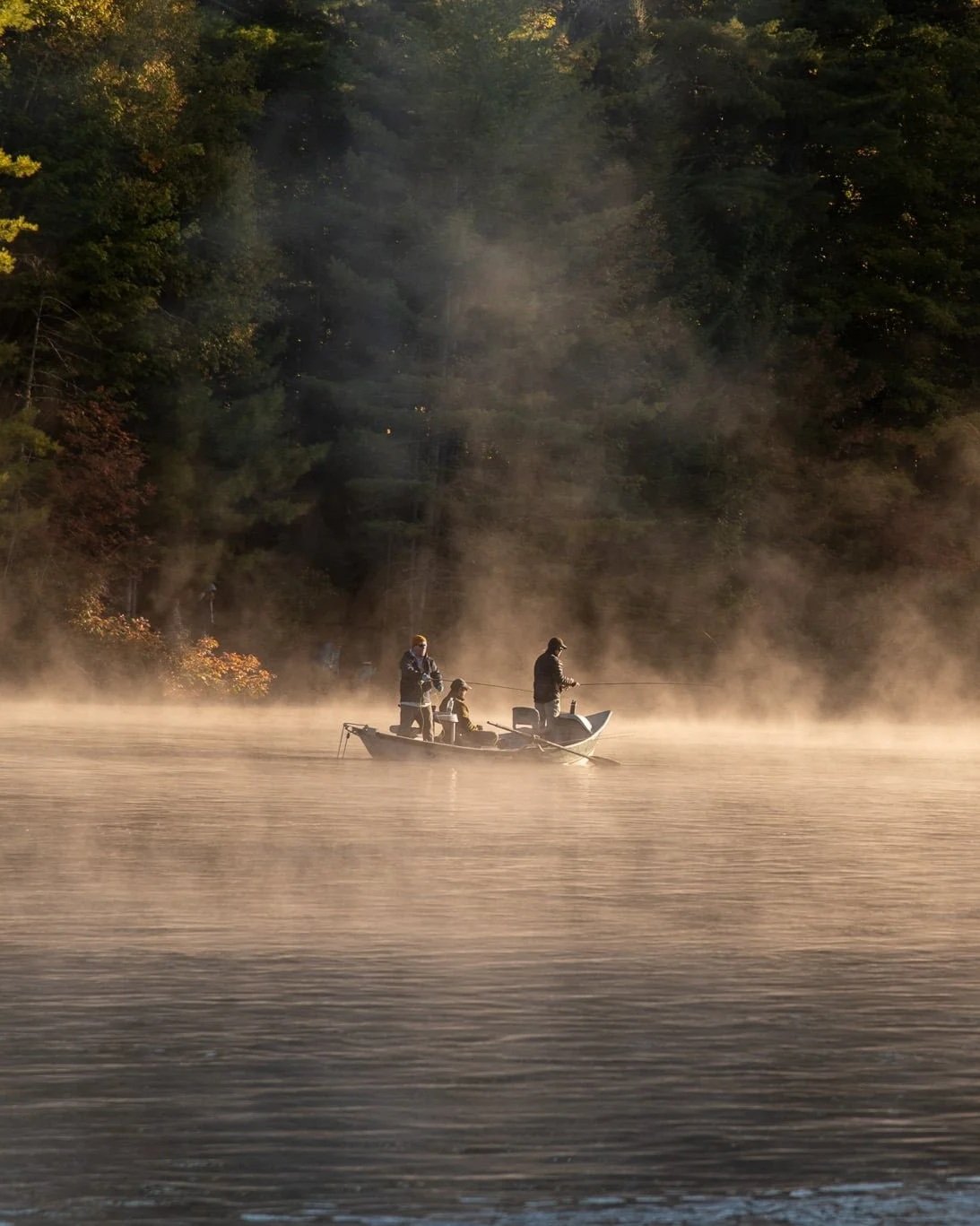 Three people fishing on a boat on a misty lake with a forested shoreline in the background.