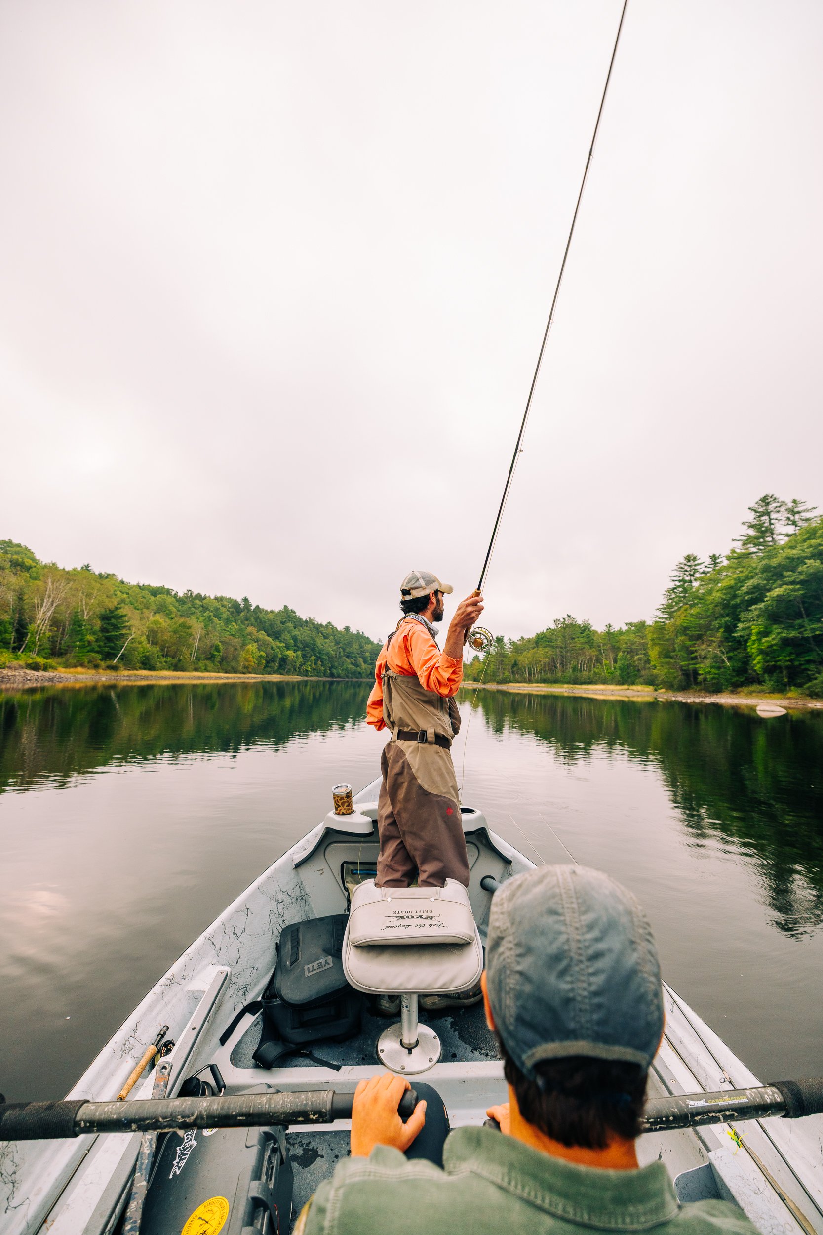 Two men fishing from a boat on a calm river surrounded by trees, overcast sky.