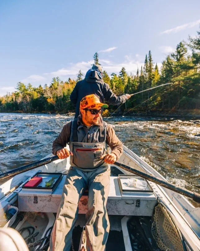 Two men fishing from a boat on a river surrounded by trees, with clear blue sky above.