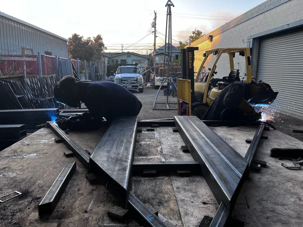 Two workers welding steel beams on a flatbed in an outdoor industrial area during sunset.