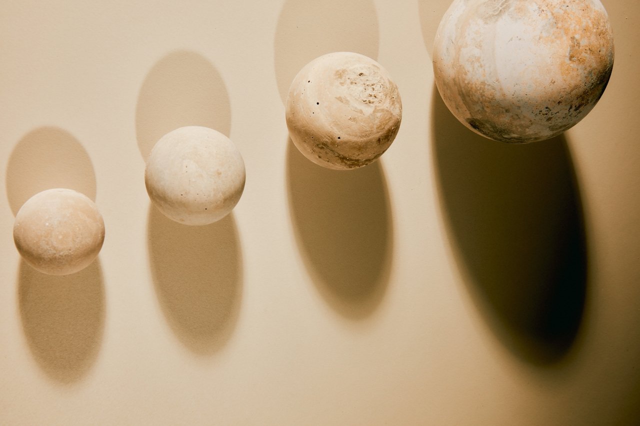Four spherical objects resembling planets or stones, arranged from smallest to largest, casting shadows on a beige surface.