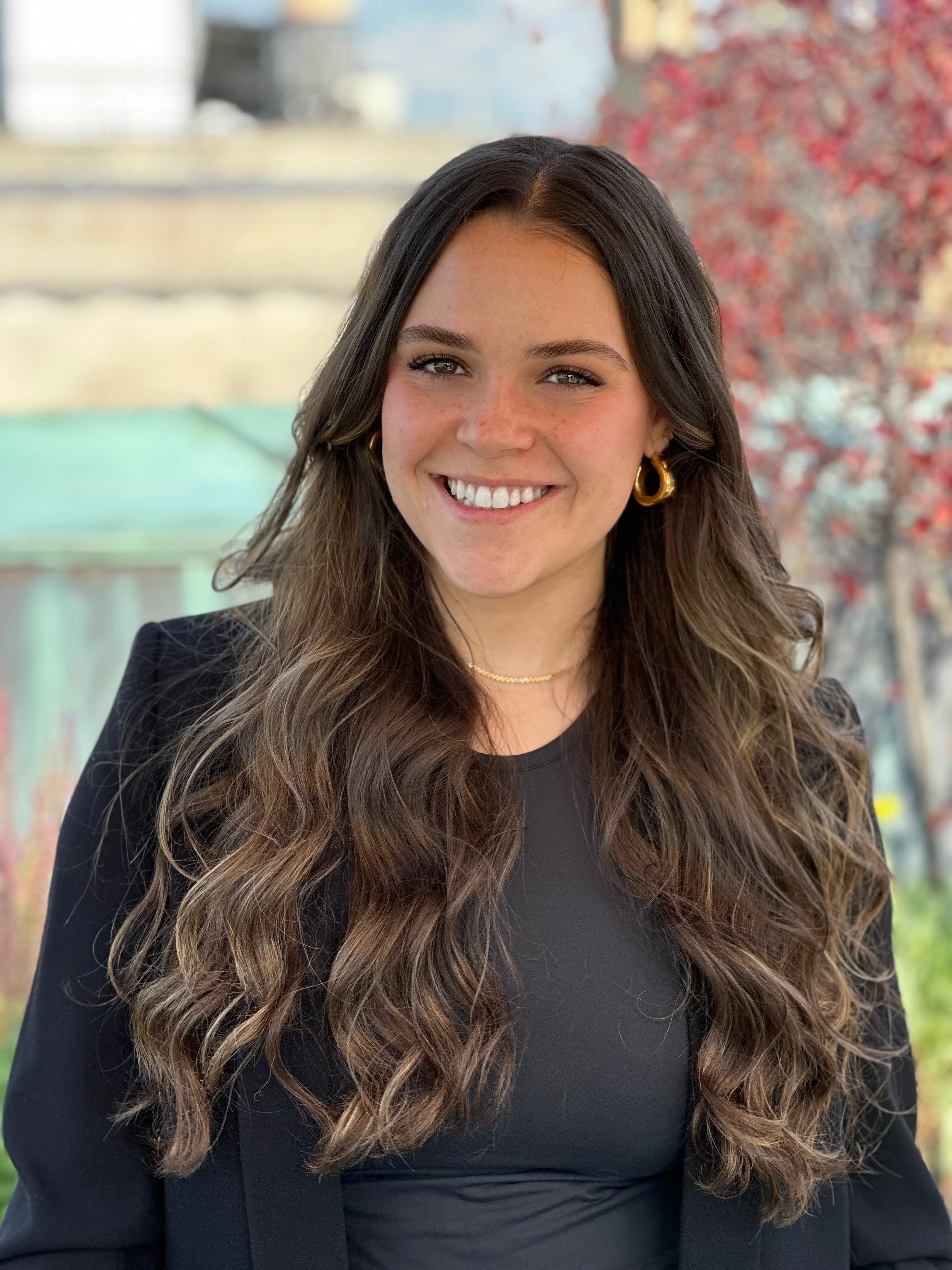 Young woman with long wavy brown hair, wearing gold hoop earrings, a black top, and a black blazer, smiling outdoors with blurred trees and buildings in the background.