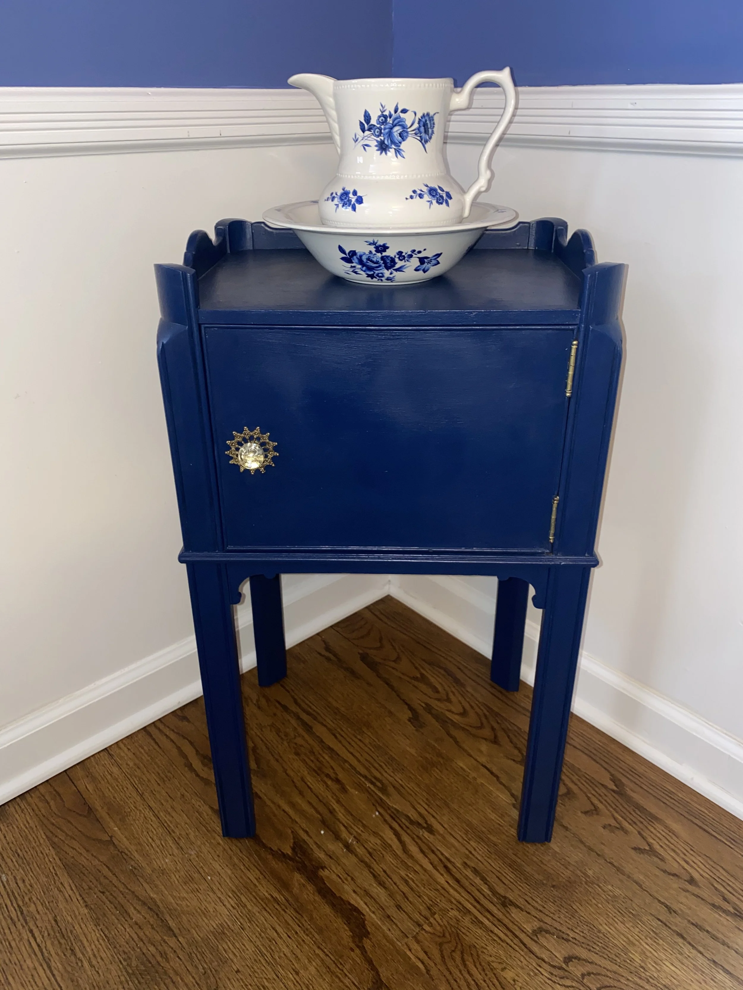 A blue wooden corner cabinet with a decorative gold knob on a hardwood floor, with a white porcelain cream pitcher and bowl decorated with blue floral patterns on top of it. The background includes a white wall with white and blue trim.