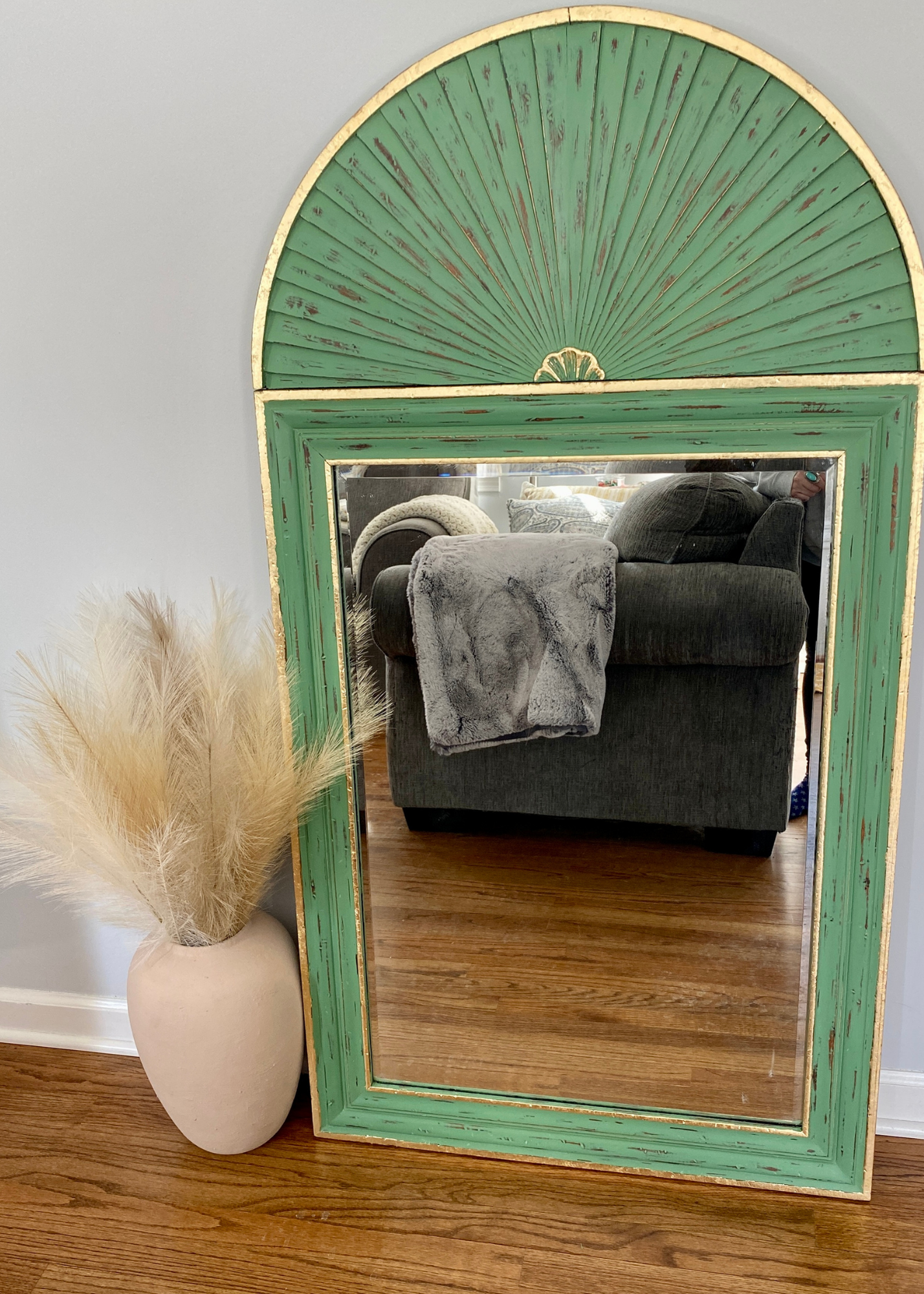 A tall, rectangular mirror with a weathered, green-painted wooden frame and a semi-circular top section. The mirror reflects a living room with a dark green armchair draped with a gray blanket, and a beige cushioned chair. On the left side of the ima