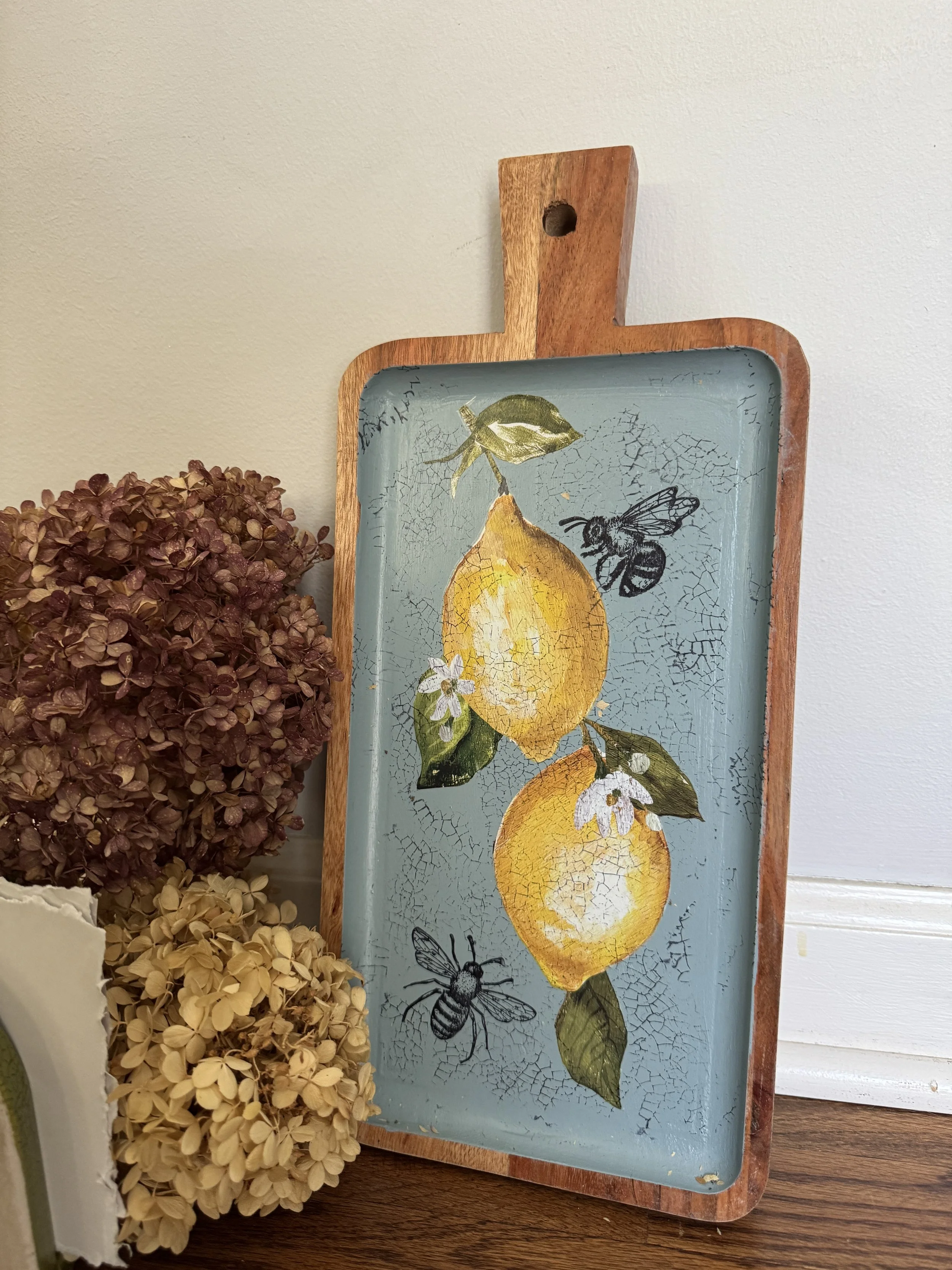 Decorative tray with painted lemons, leaves, and bees, placed beside dried hydrangea flowers on a wooden surface.