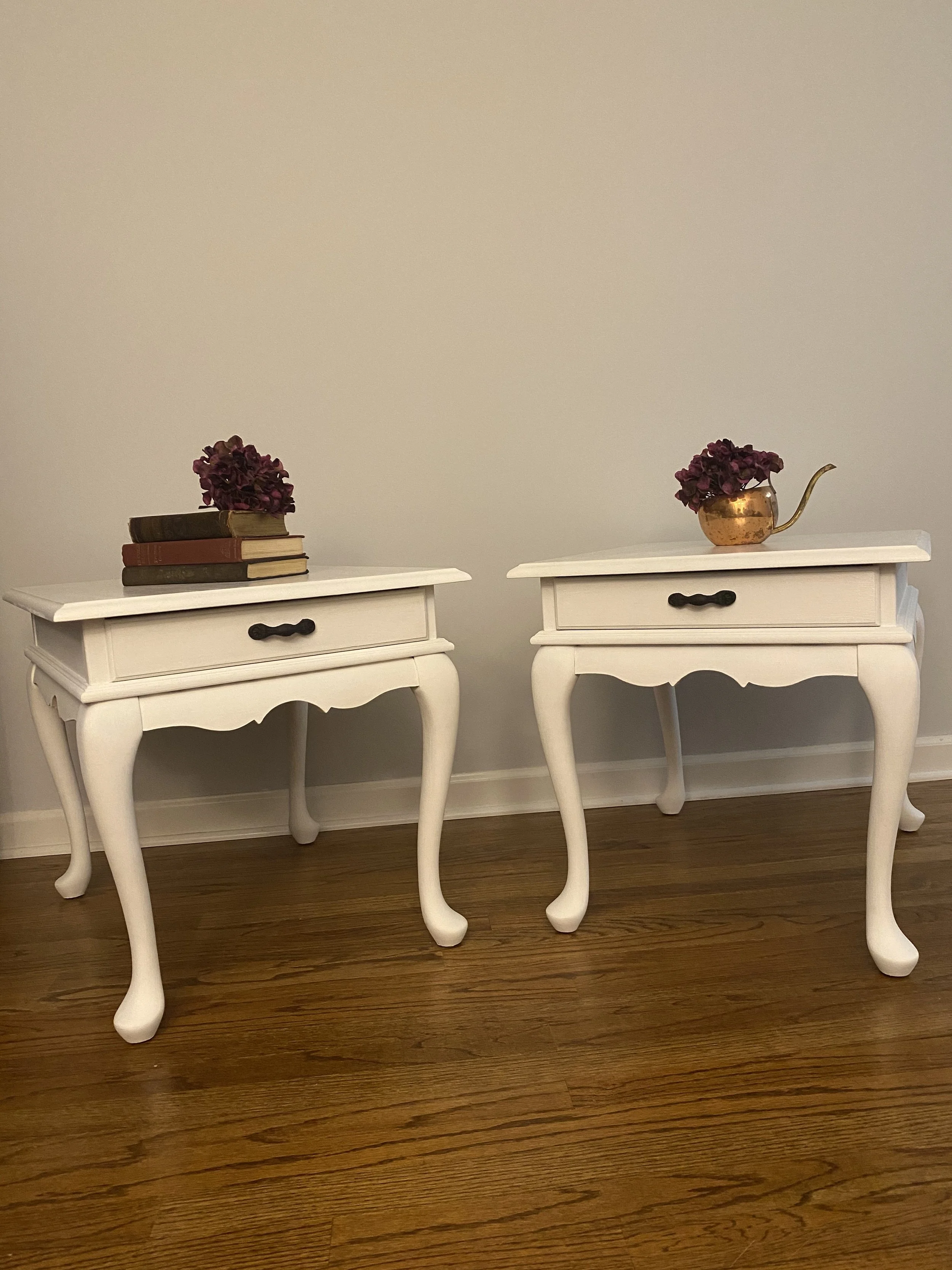 Two white vintage-style side tables with black handles, each topped with a small bouquet of dark purple flowers in a copper watering can, on a hardwood floor against a plain beige wall.