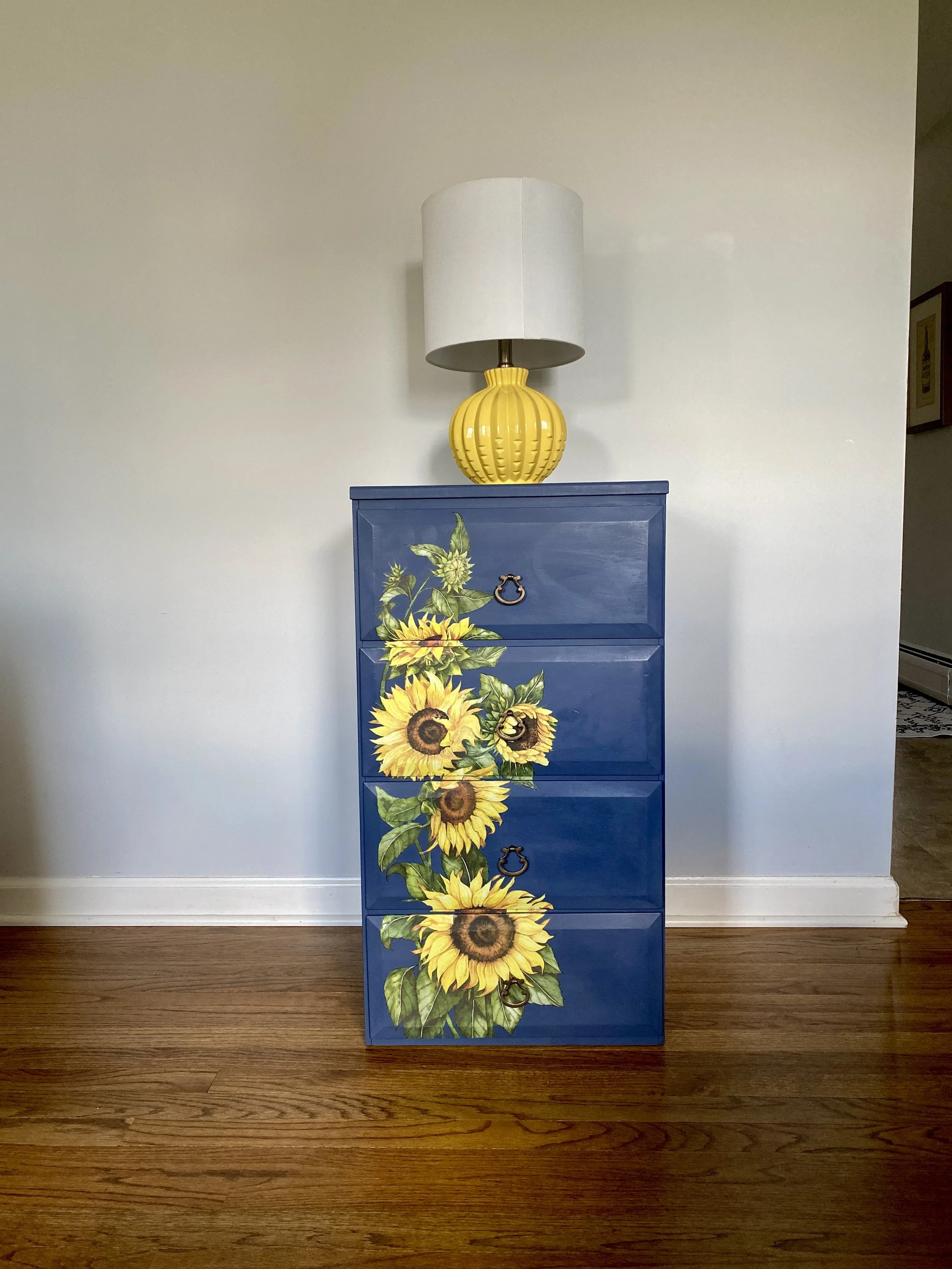 Blue dresser with sunflower design on the front, yellow ceramic lamp on top, white wall background, wooden floor.