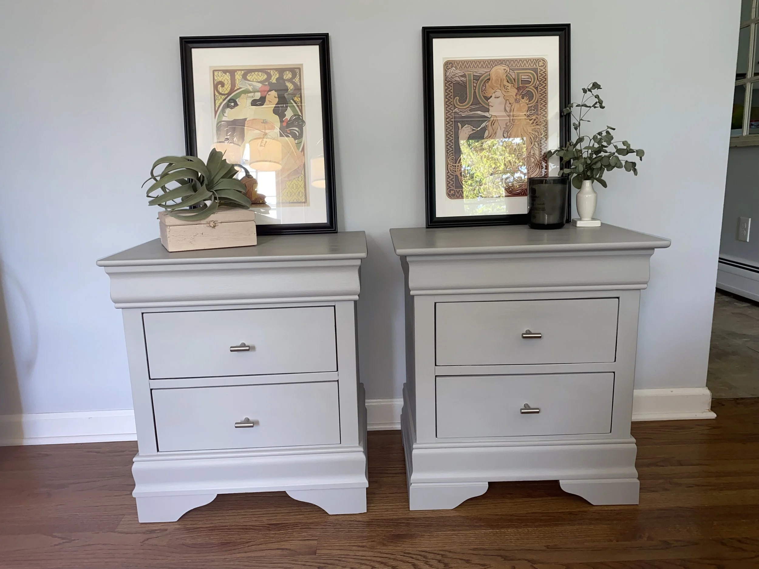 Two white dressers with framed artwork on top, each decorated with a potted plant, placed against a light-colored wall.