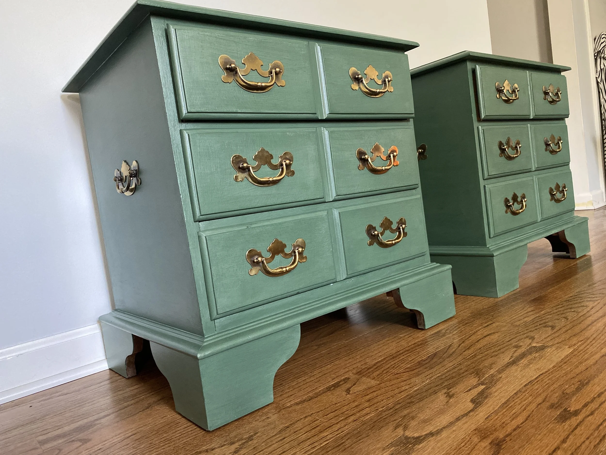 Two green wooden dressers with brass handles placed on a wooden floor against a light-colored wall.