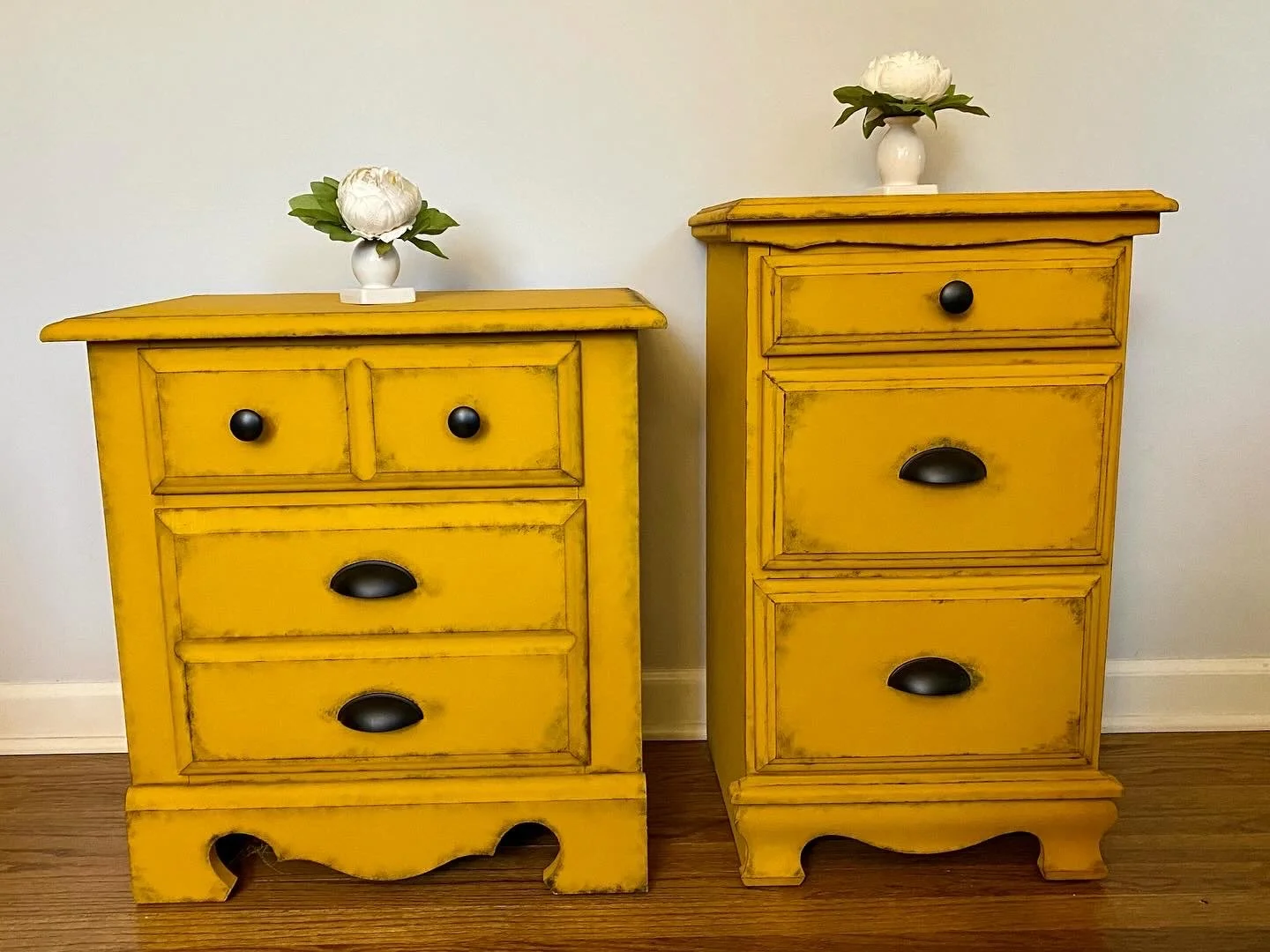 Two matching yellow wooden nightstands with distressed finish, each with three drawers and black handles, placed against a white wall, with white flower vases with large white flowers on top.