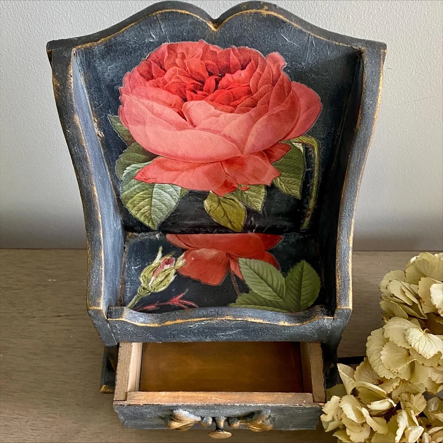 Decorative wooden box with a painted pink rose and green leaves on the inside lid, situated on a wooden surface next to a cluster of dried white flowers.