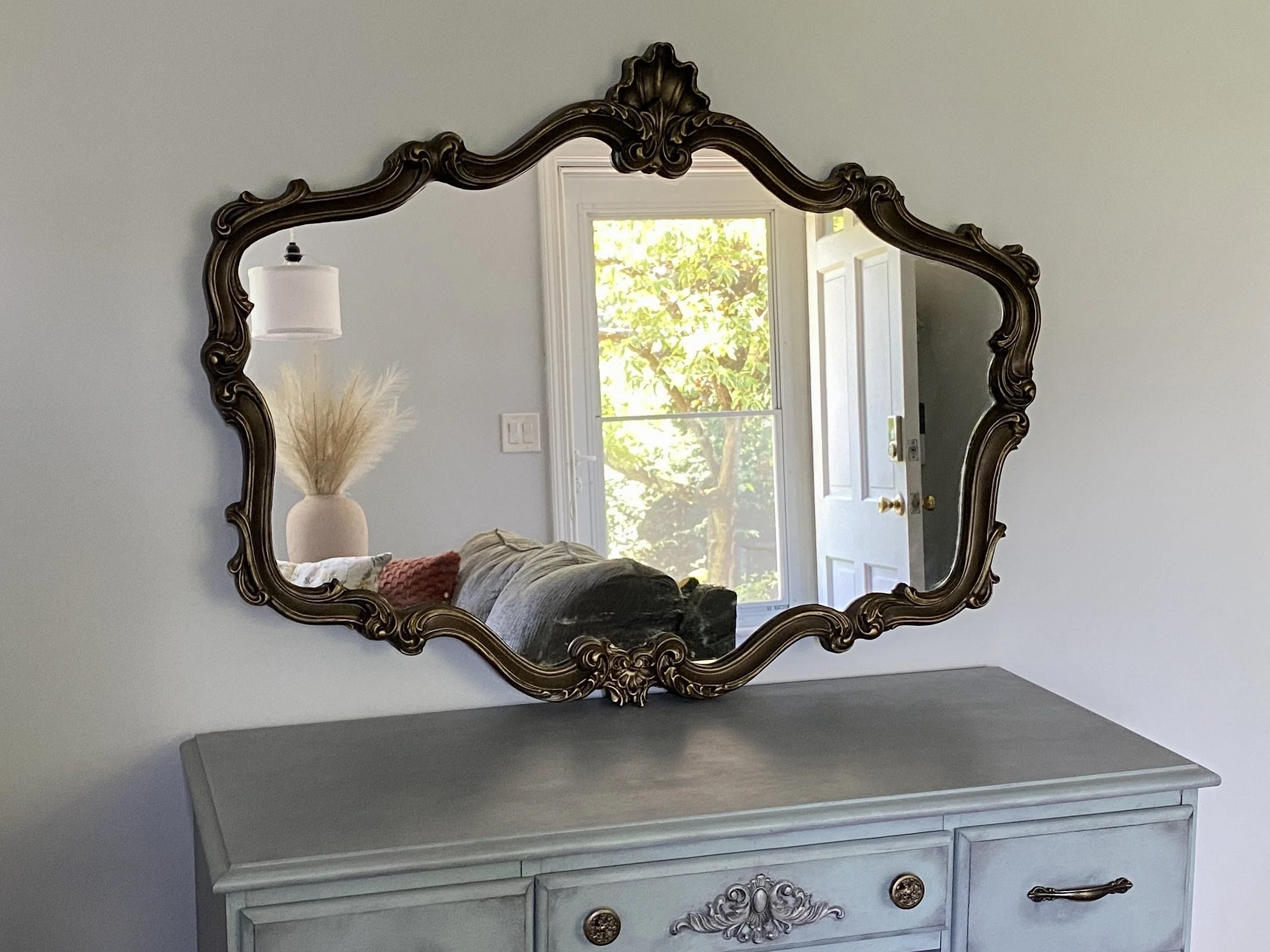 A decorative antique-style mirror with a dark ornate frame hanging on a light colored wall. The reflection in the mirror shows a white door with a window revealing trees outside, and part of a white dresser below the mirror.